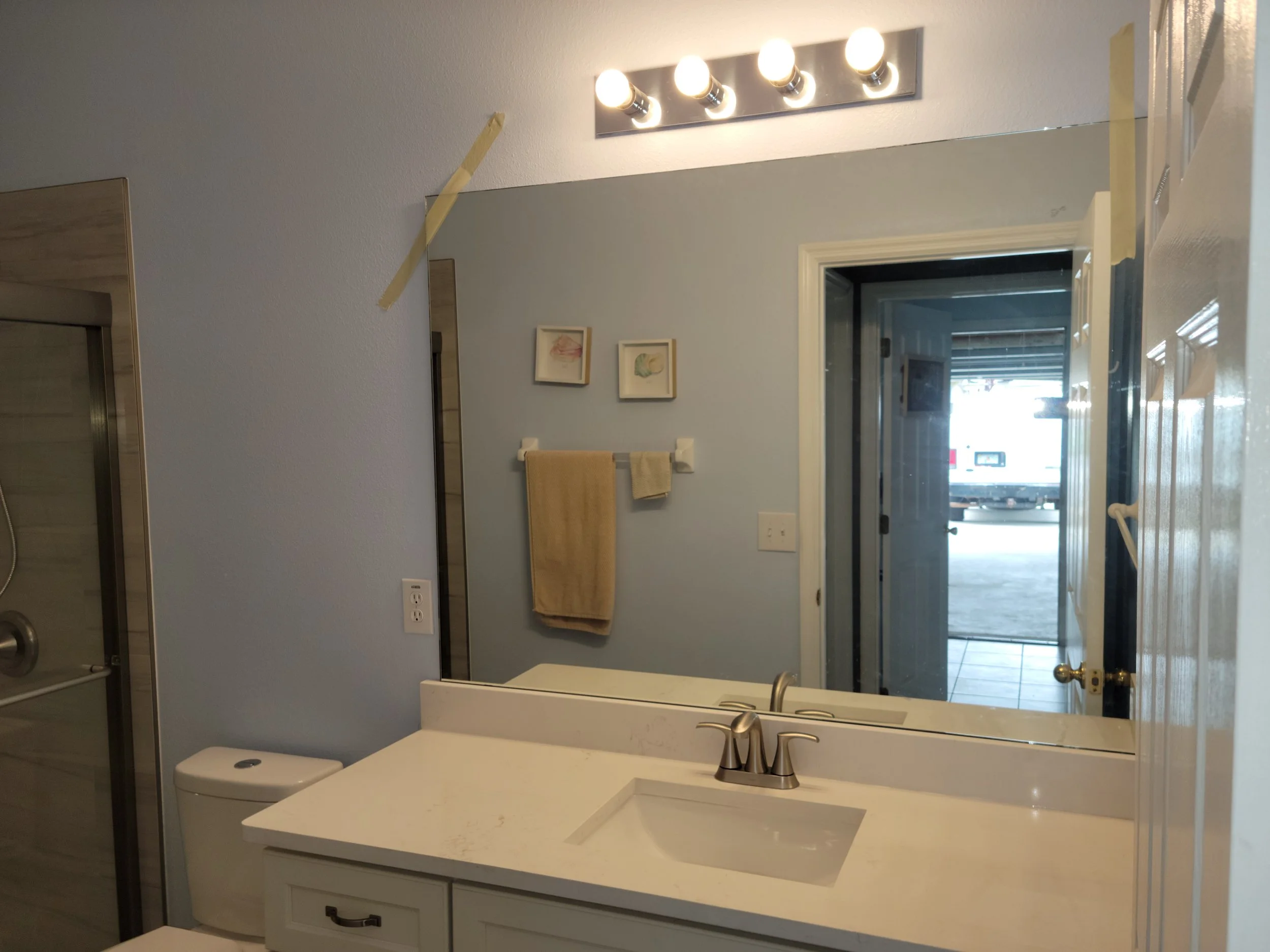 Bathroom mirror with four light bulbs, countertop with sink, towel rack with yellow towel, artwork on wall, door open to outside, and partially visible shower.