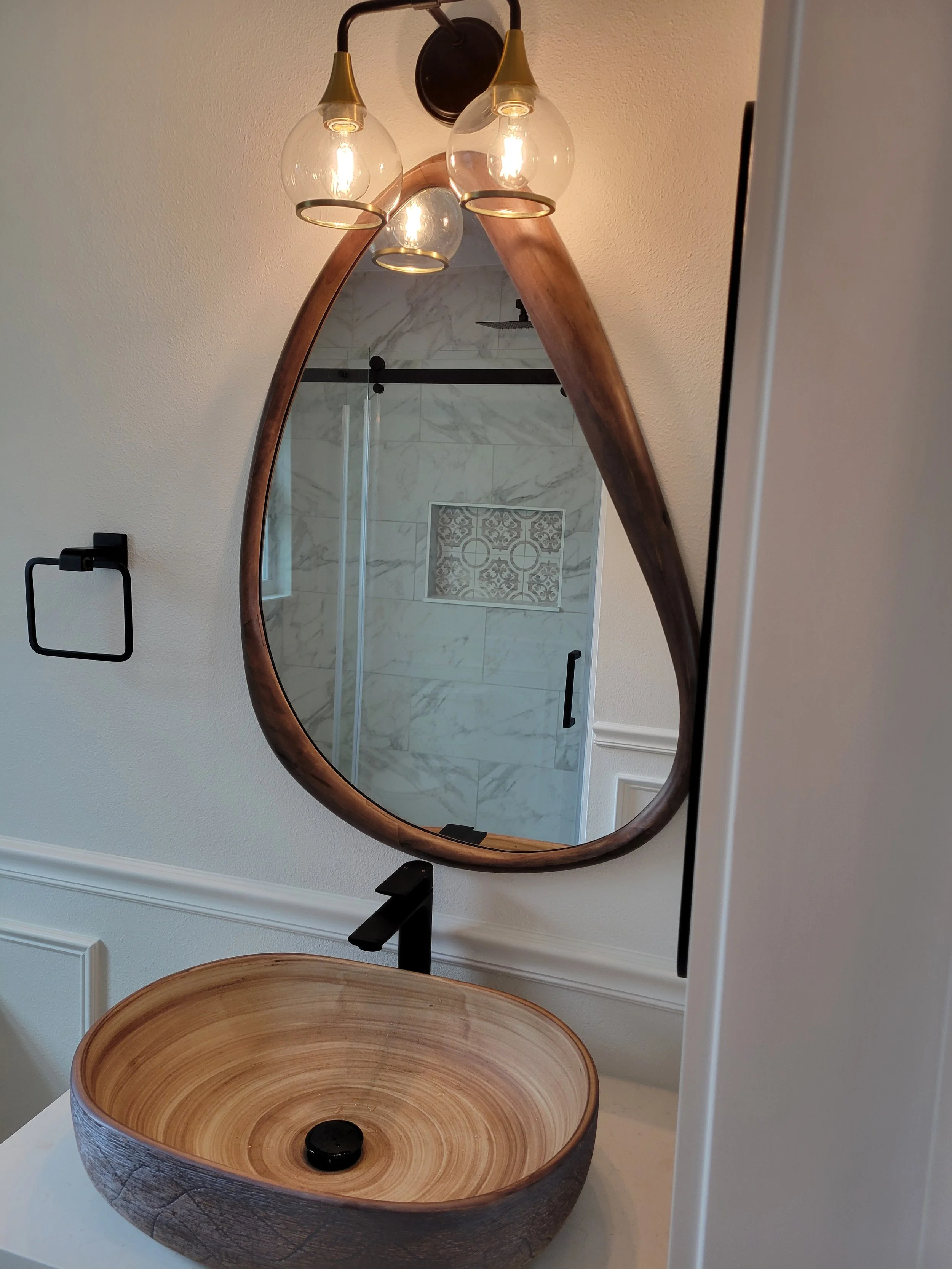 Bathroom vanity with a wooden vessel sink equipped with a black faucet, and teardrop-shaped mirror with a wooden frame