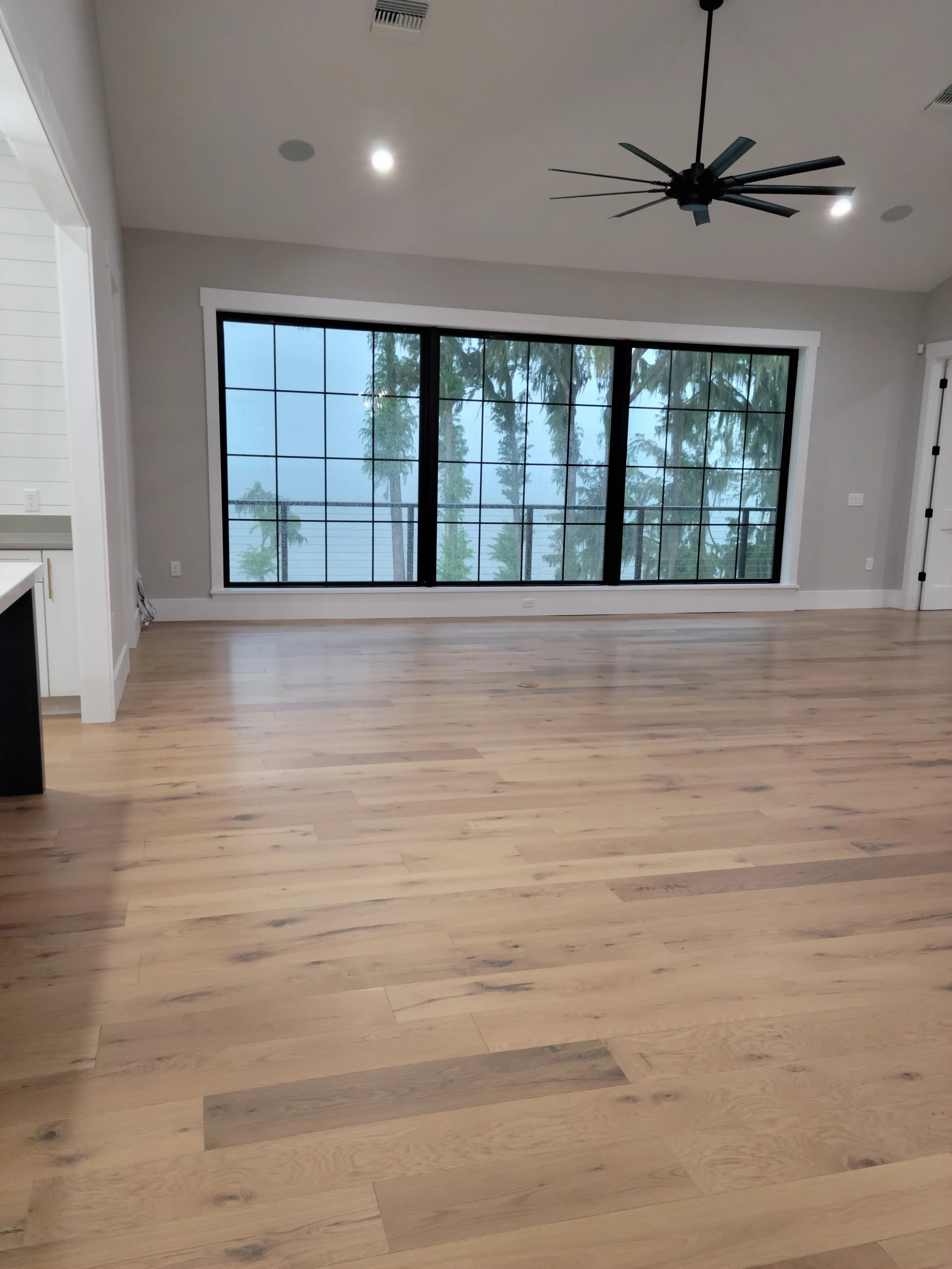 Empty living room with light wood flooring, large window showing trees outside, ceiling fan, and recessed lighting.