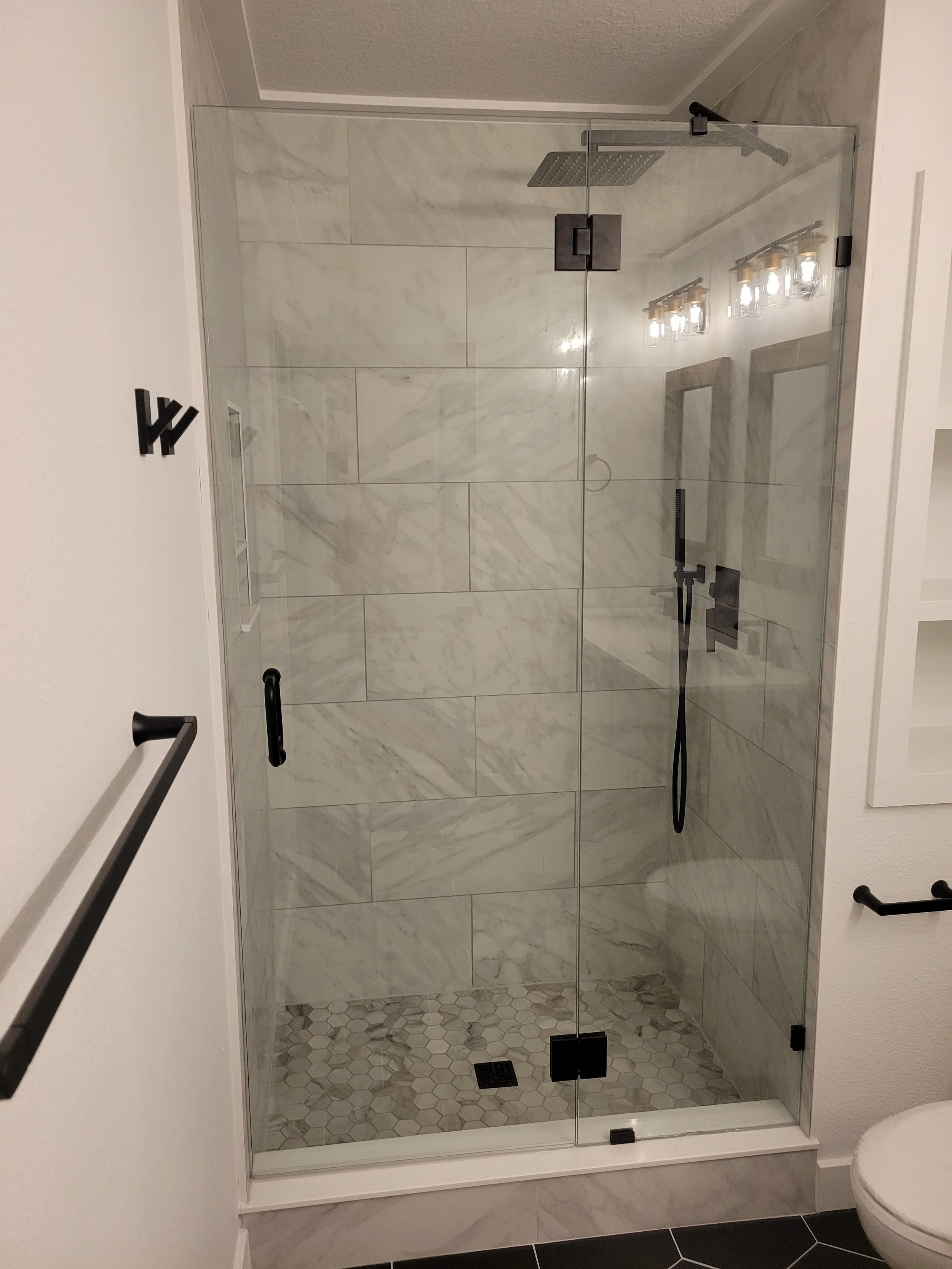 A modern walk-in shower with glass doors, white marble tiles, and black fixtures, including a rainfall showerhead and hand shower, in a contemporary bathroom.