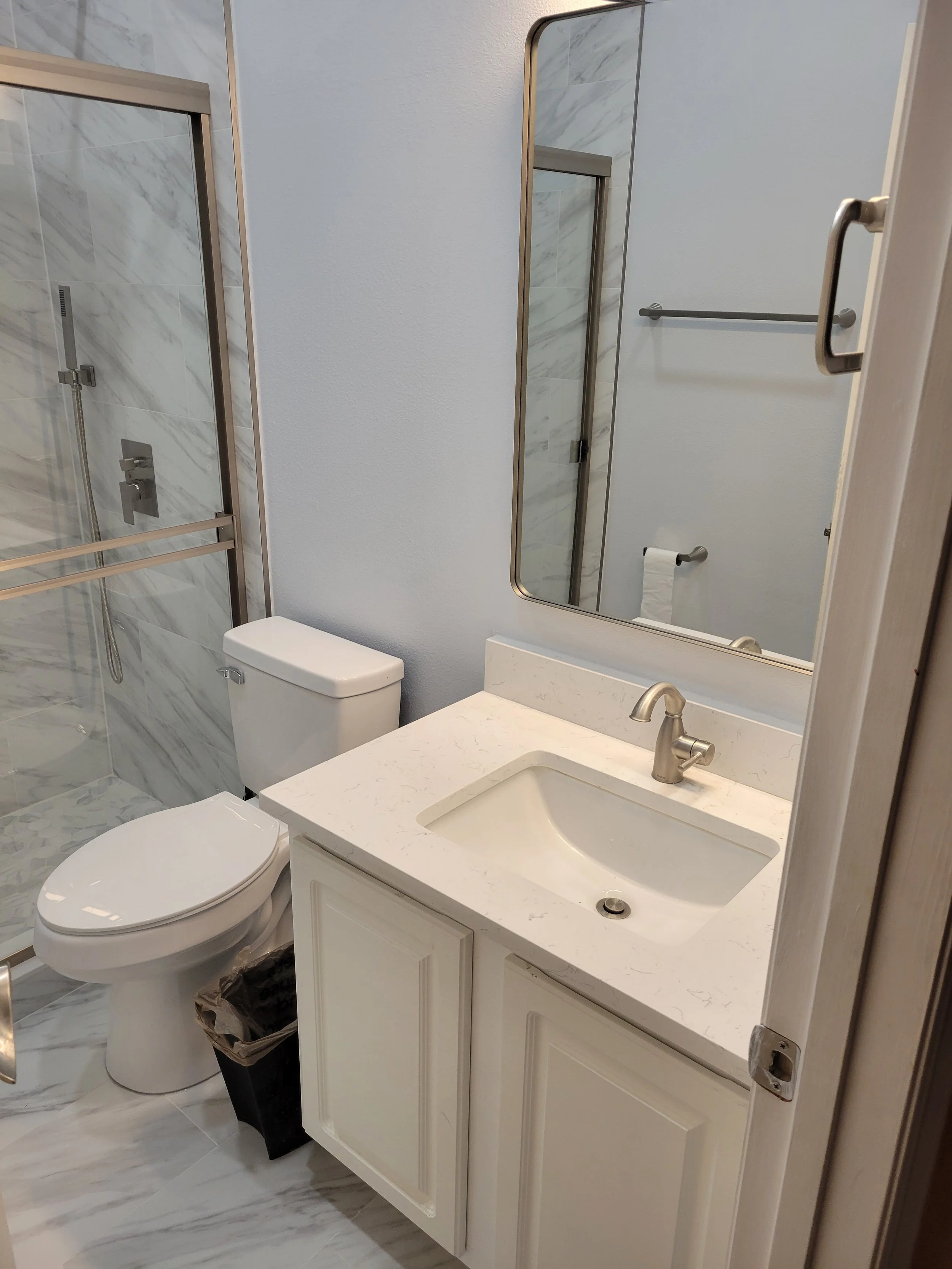 Bathroom with a white vanity, sink, mirror, toilet, and a glass-enclosed shower with marble tiles.