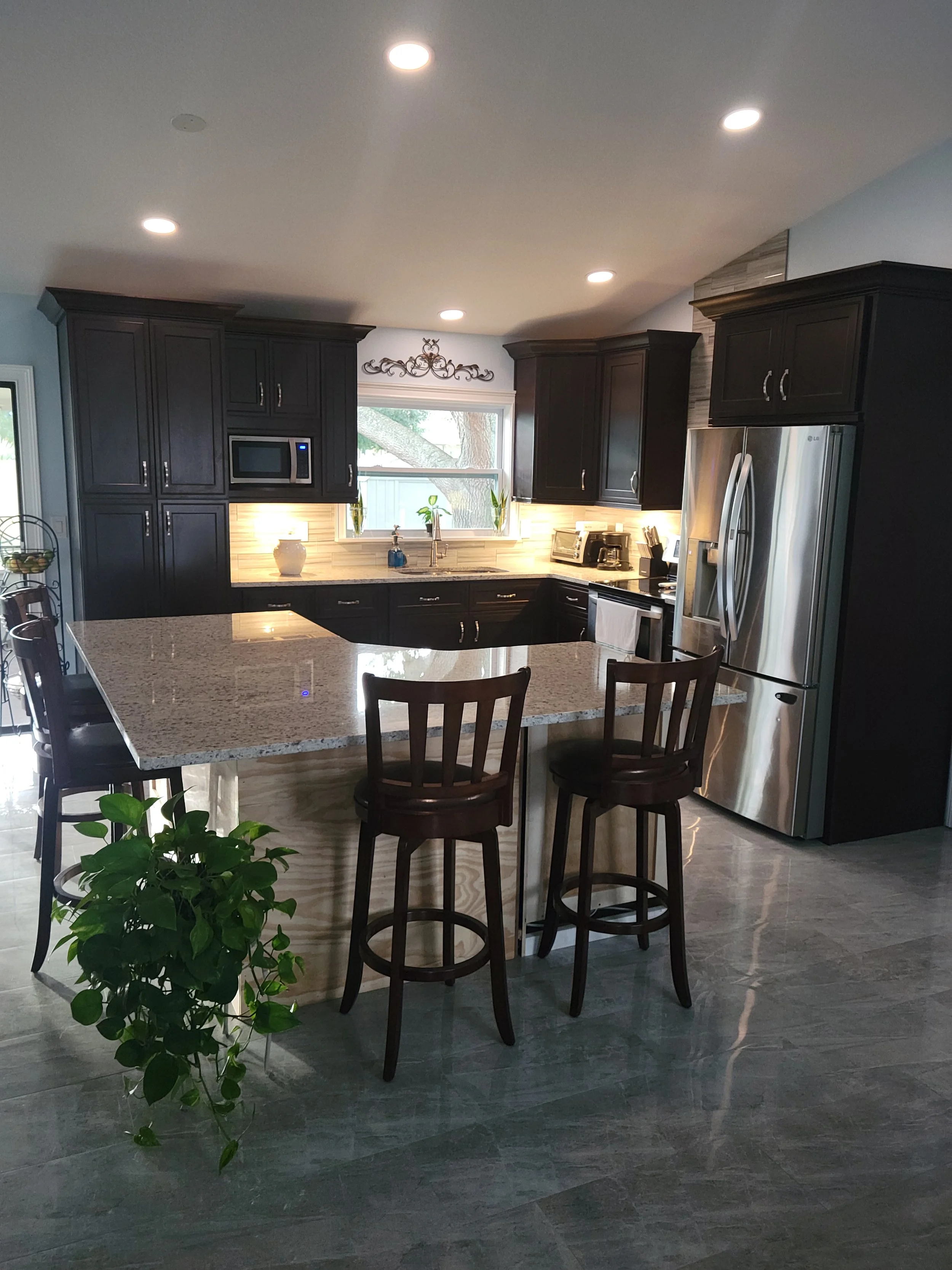 Modern kitchen with black cabinets, stainless steel refrigerator, granite island with two wooden bar stools, and a window overlooking outdoor trees.