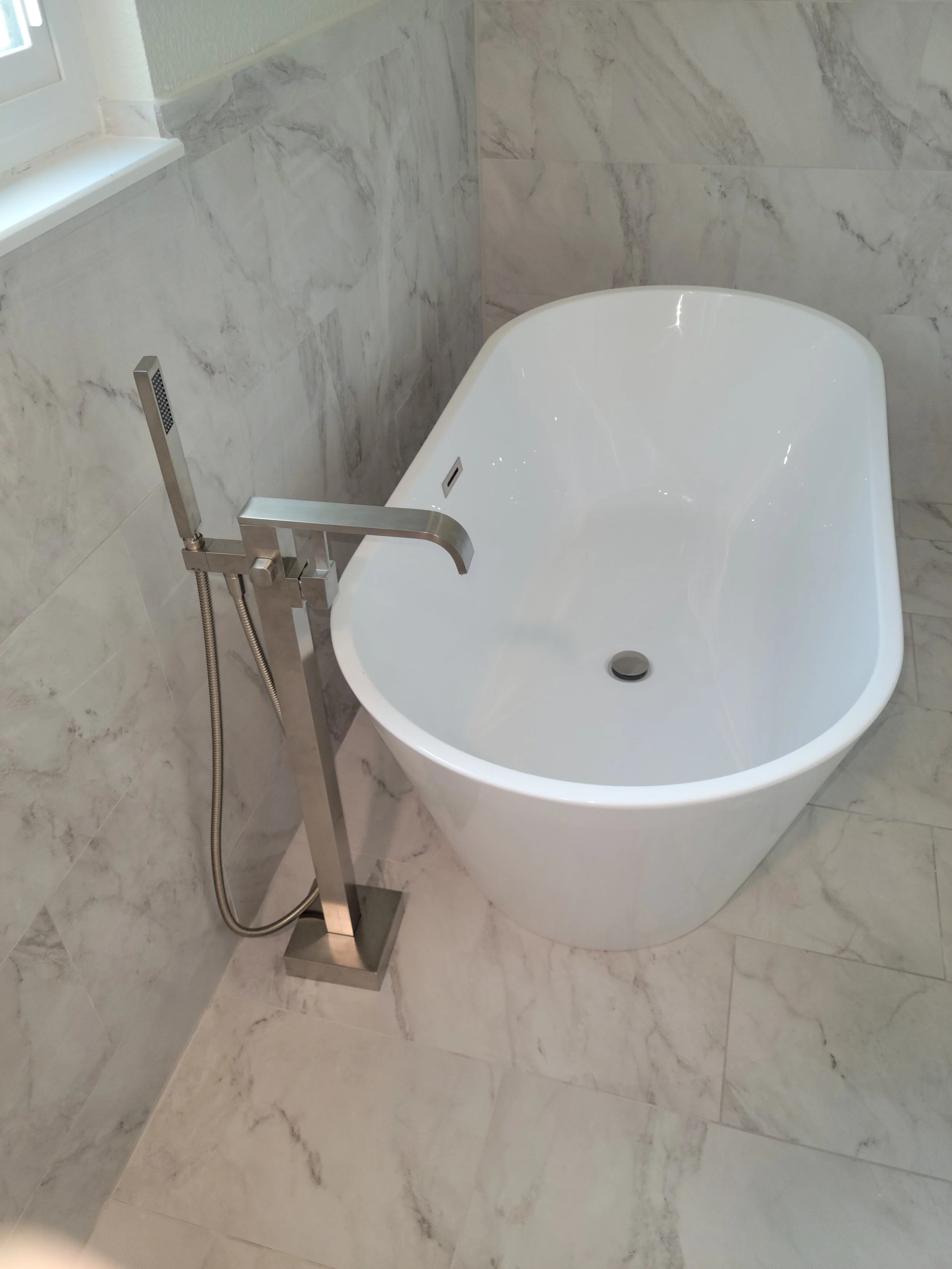 Modern freestanding bathtub with chrome faucet and hand shower in a marble-tiled bathroom.