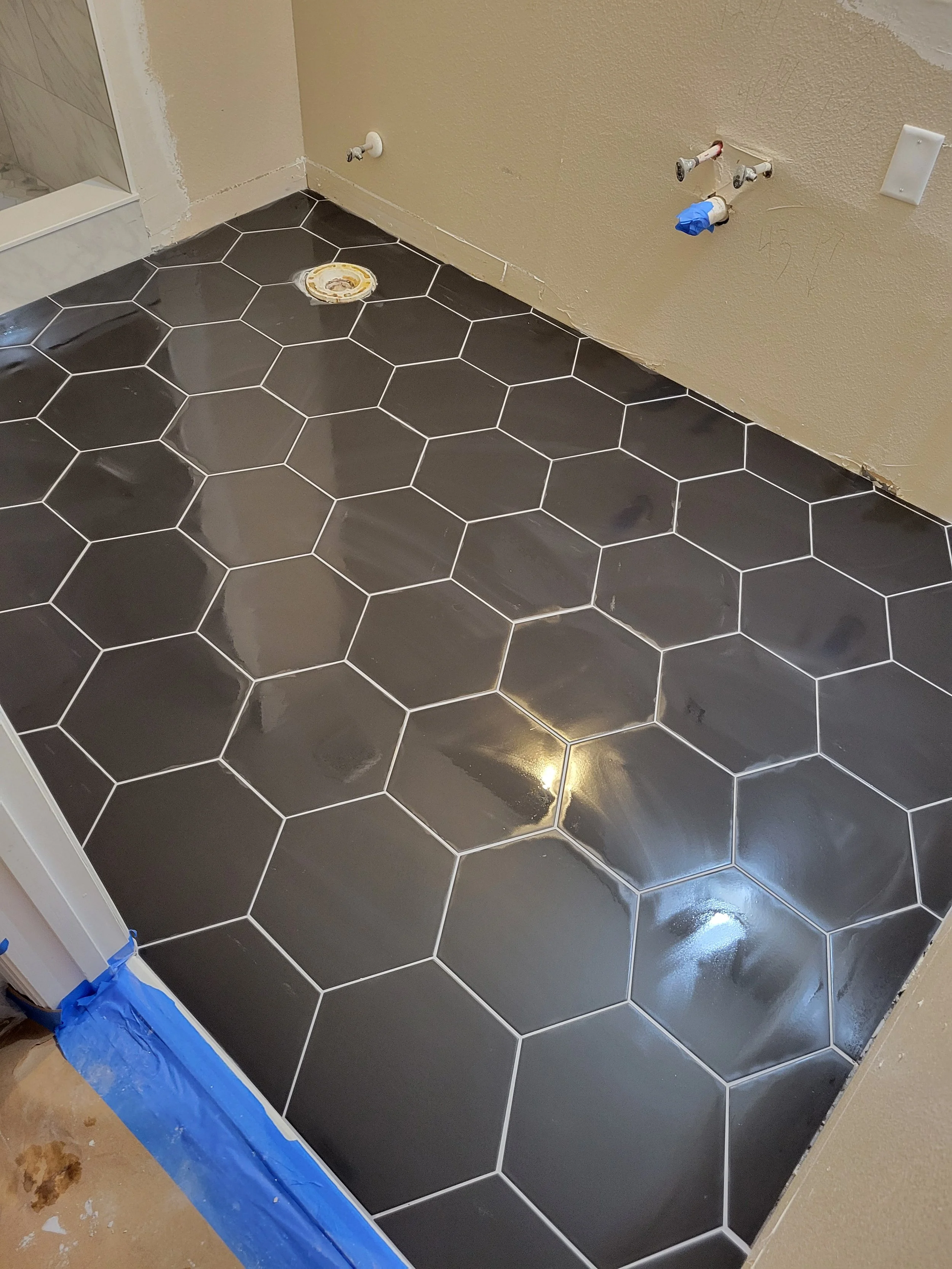 Bathroom floor with black hexagonal tiles that are still wet, with some construction tools and materials visible, including a pre-existing drain and exposed plumbing connections.