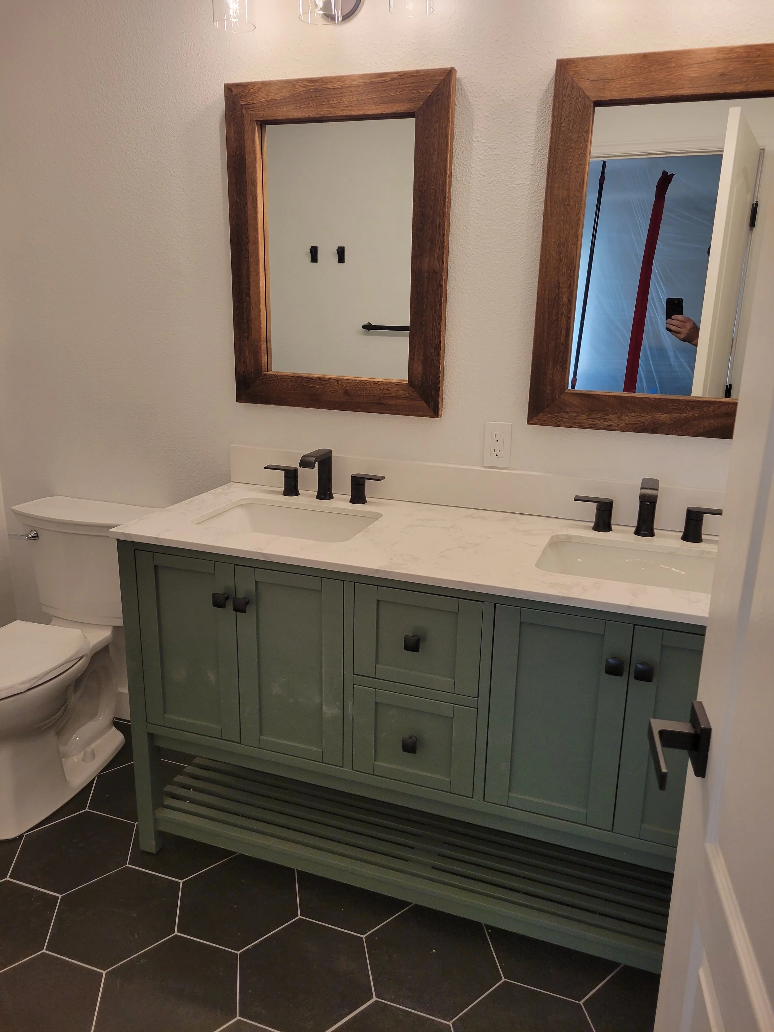 A modern bathroom vanity with a marble countertop, black fixtures, and a green cabinet, with two mirrors framed in wood above the sinks.