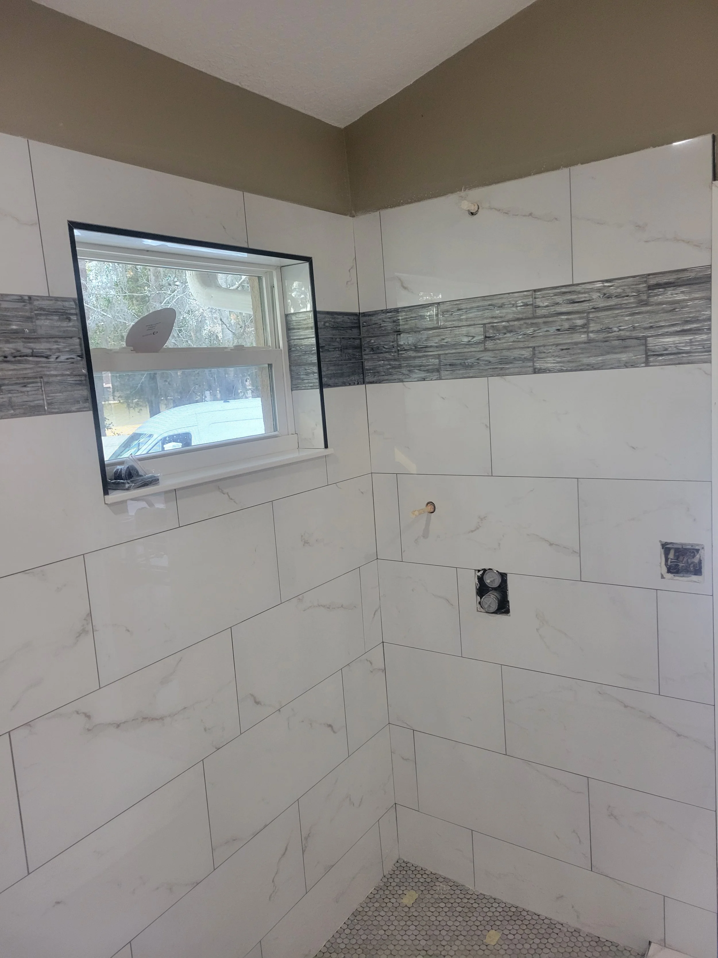 A partially finished bathroom shower with large white marble tiles and a horizontal accent strip of gray and white tiles, and a small window.