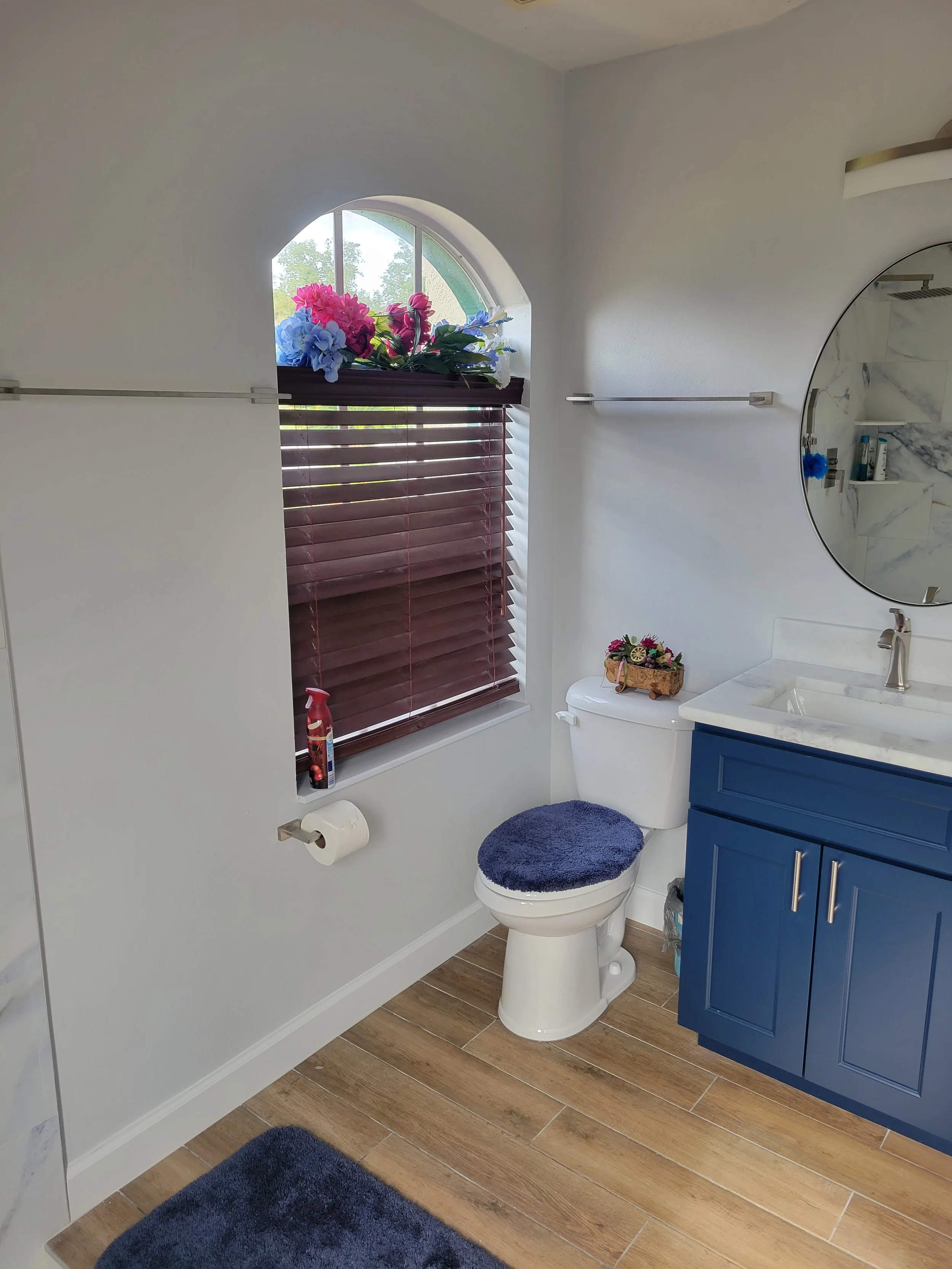Bathroom with blue vanity, white toilet with a blue fuzzy seat cover, window with brown blinds, artificial flowers on the window ledge, and a round mirror above the sink.