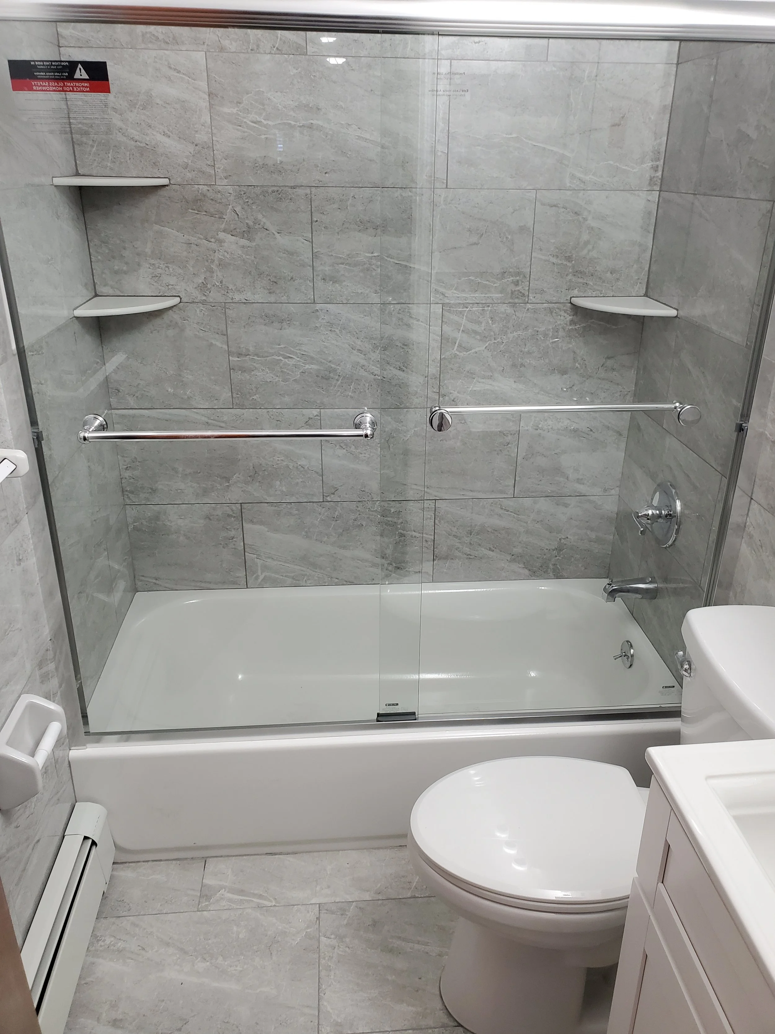 Bathroom with a glass-enclosed shower featuring two white corner shelves, a grab bar, and a chrome shower fixture. There is a toilet and a white vanity sink at the front, with gray stone tile walls and floor.