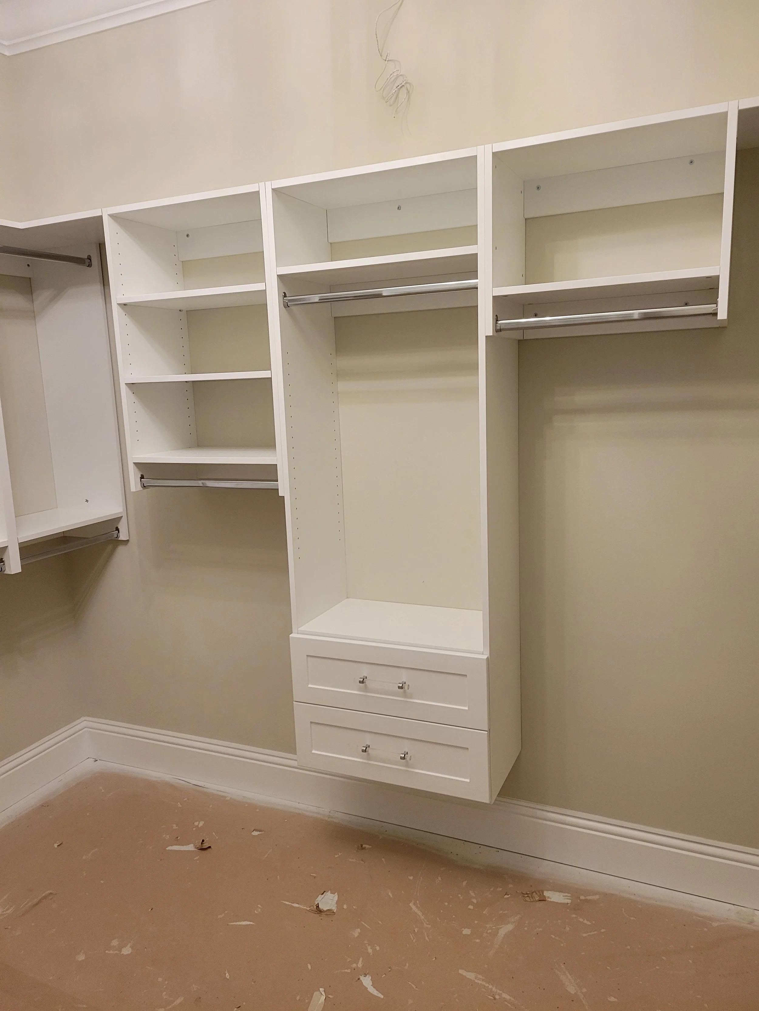 white built-in closet with shelves, drawers, and hanging rods, against beige wall, in a room with unfinished floor.