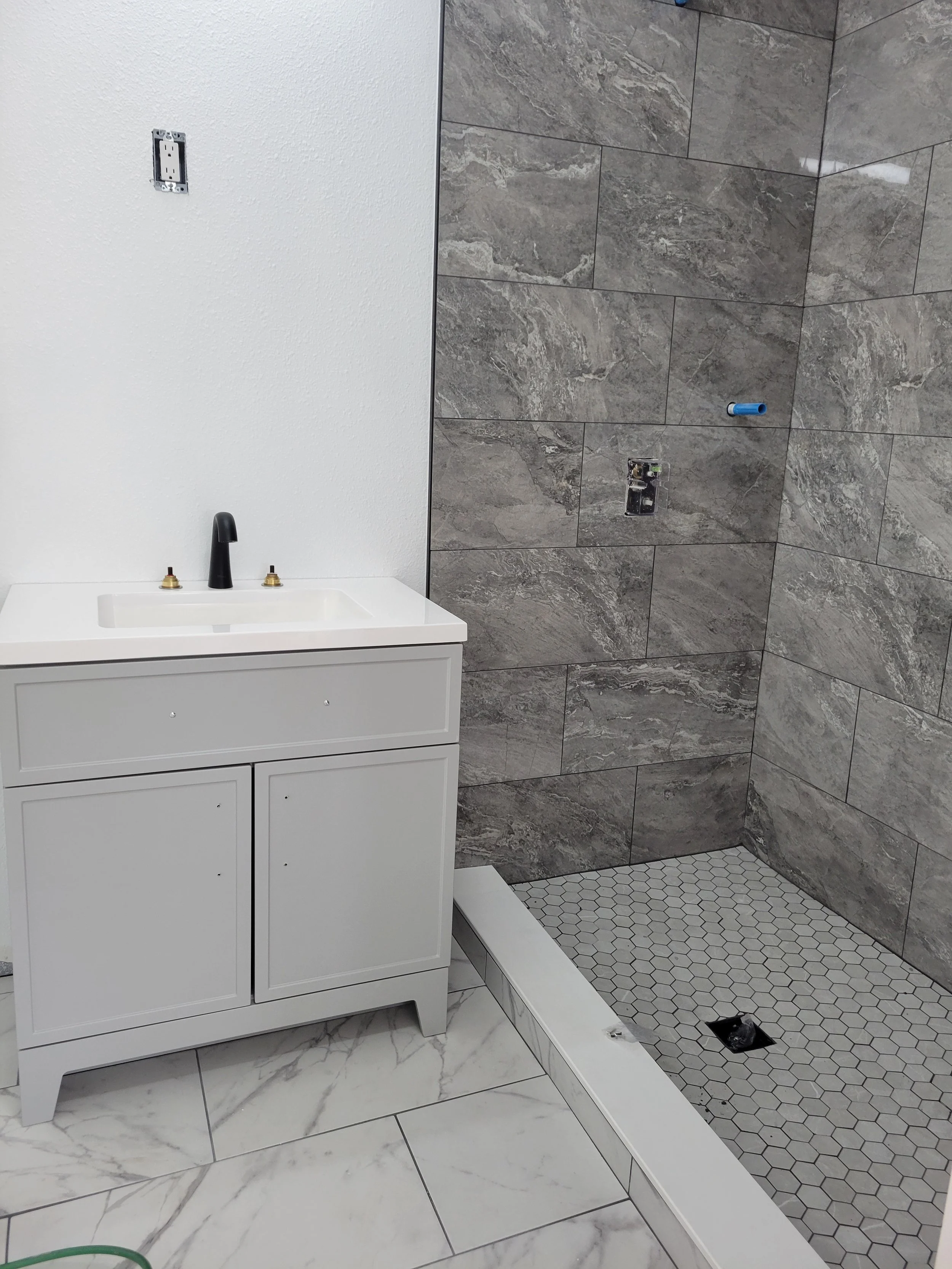 Bathroom vanity with a sink and faucet next to a walk-in shower with gray tile walls and a hexagon tile floor at the shower base, under construction.