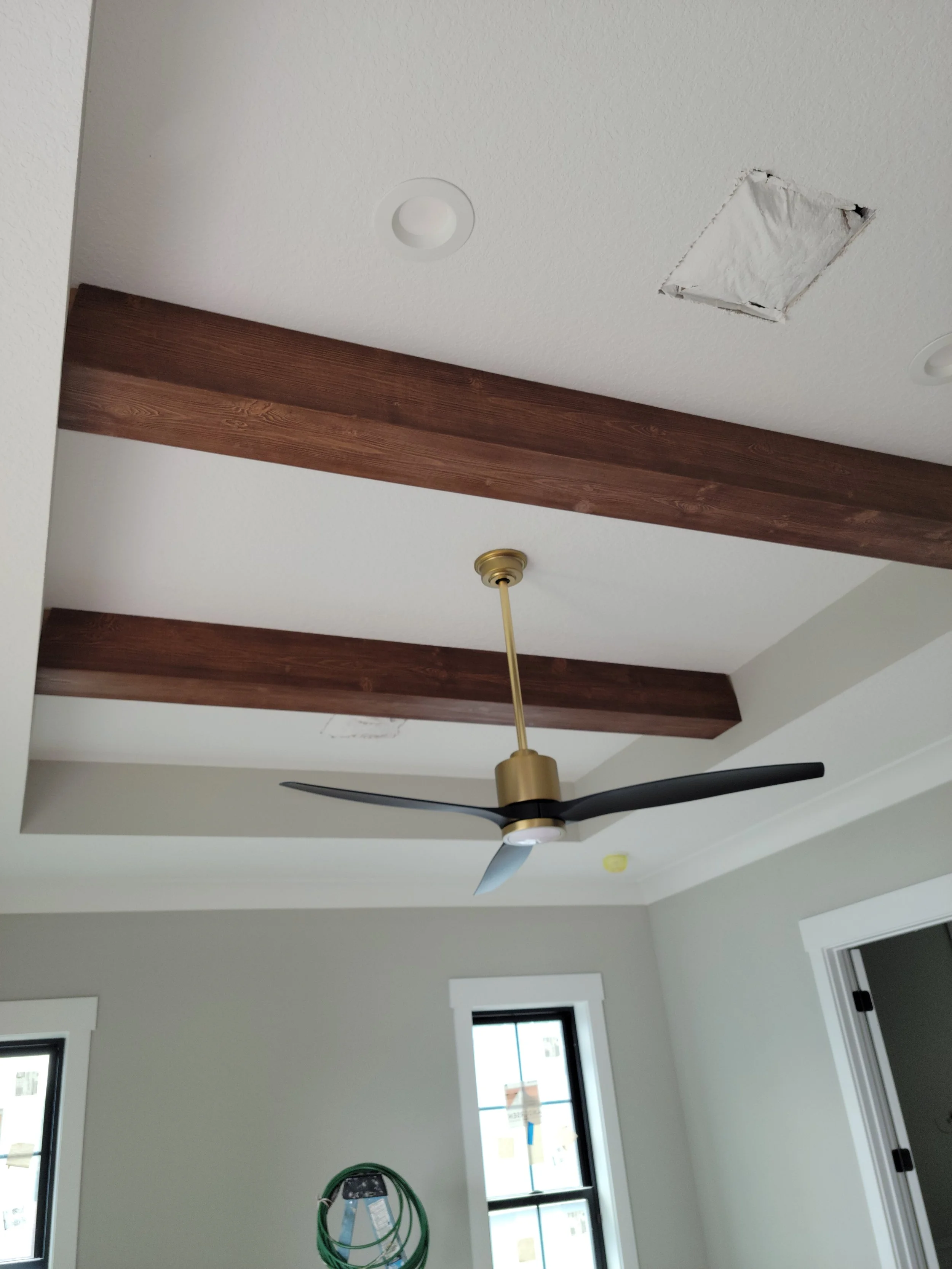 Ceiling with wooden beams, a black and gold ceiling fan, two windows, and a missing ceiling vent or fixture in a room under construction or renovation.