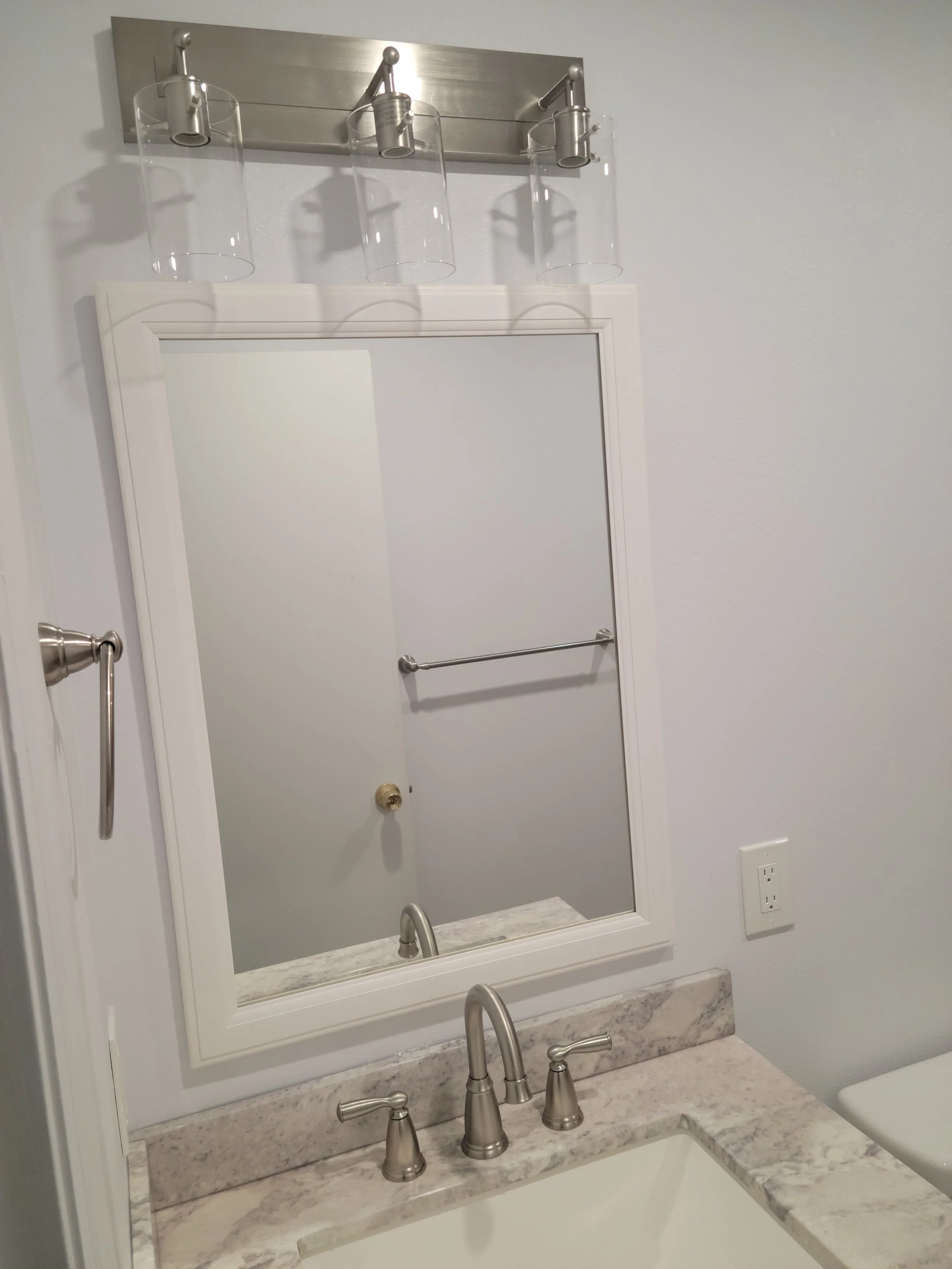 Bathroom mirror with a marble countertop and a three-light fixture above it, the mirror reflects a towel bar and a closed door.