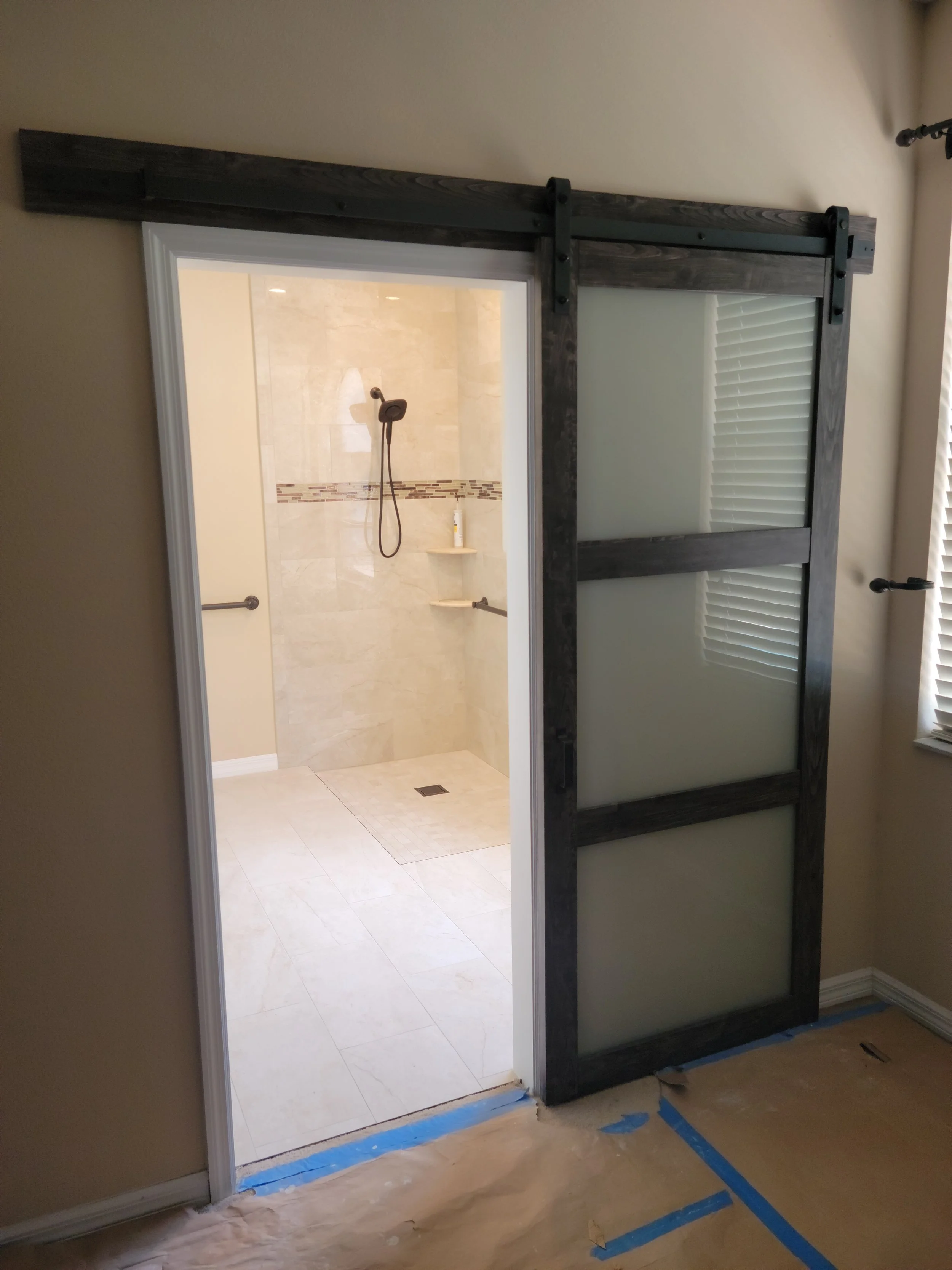 Bathroom shower with beige tiles, shower head, and grab bars, viewed through a partially open sliding door with a black wooden frame.
