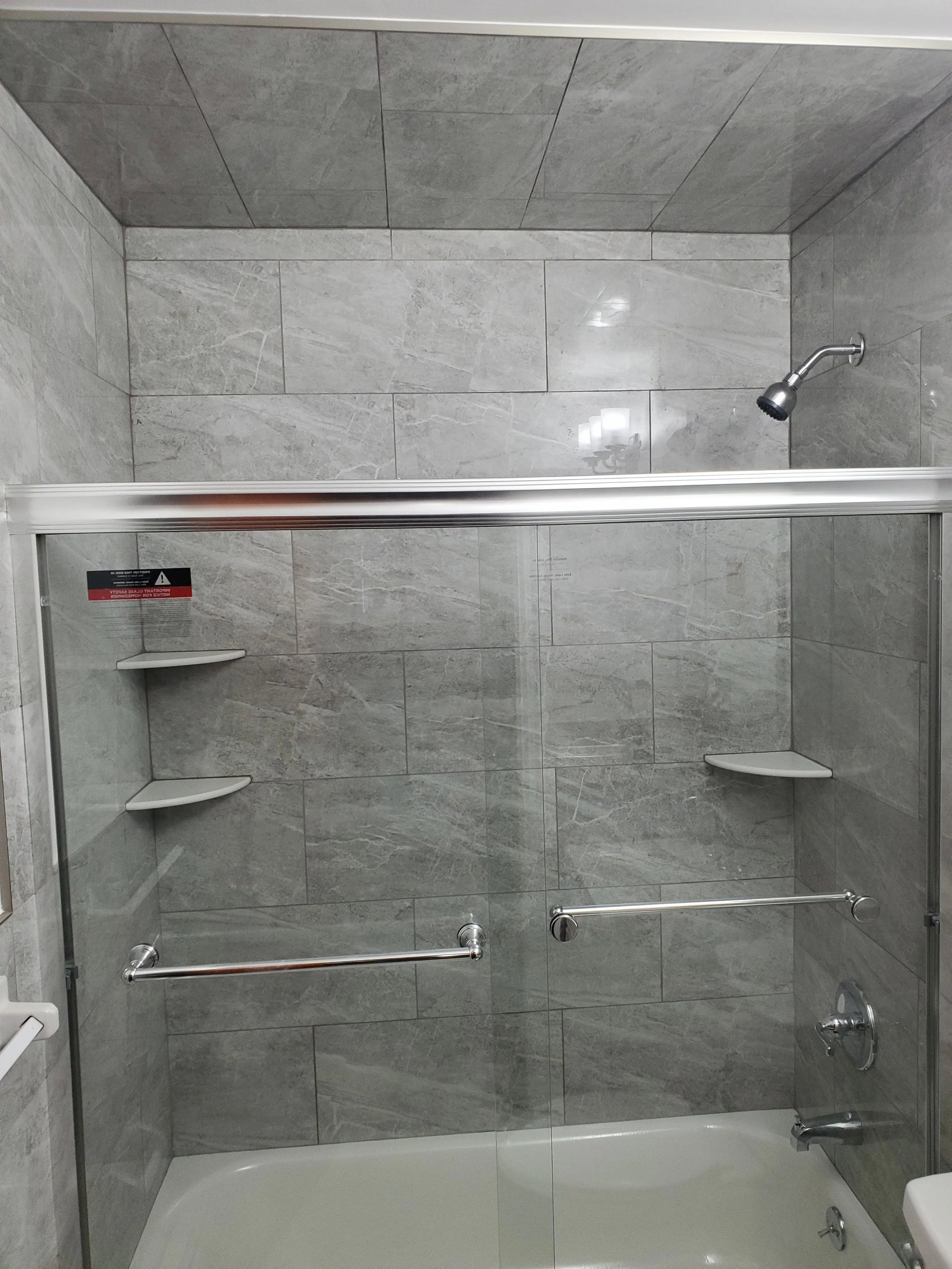 Gray tiled shower with glass door, silver showerhead, built-in corner shelves, and grab bars.
