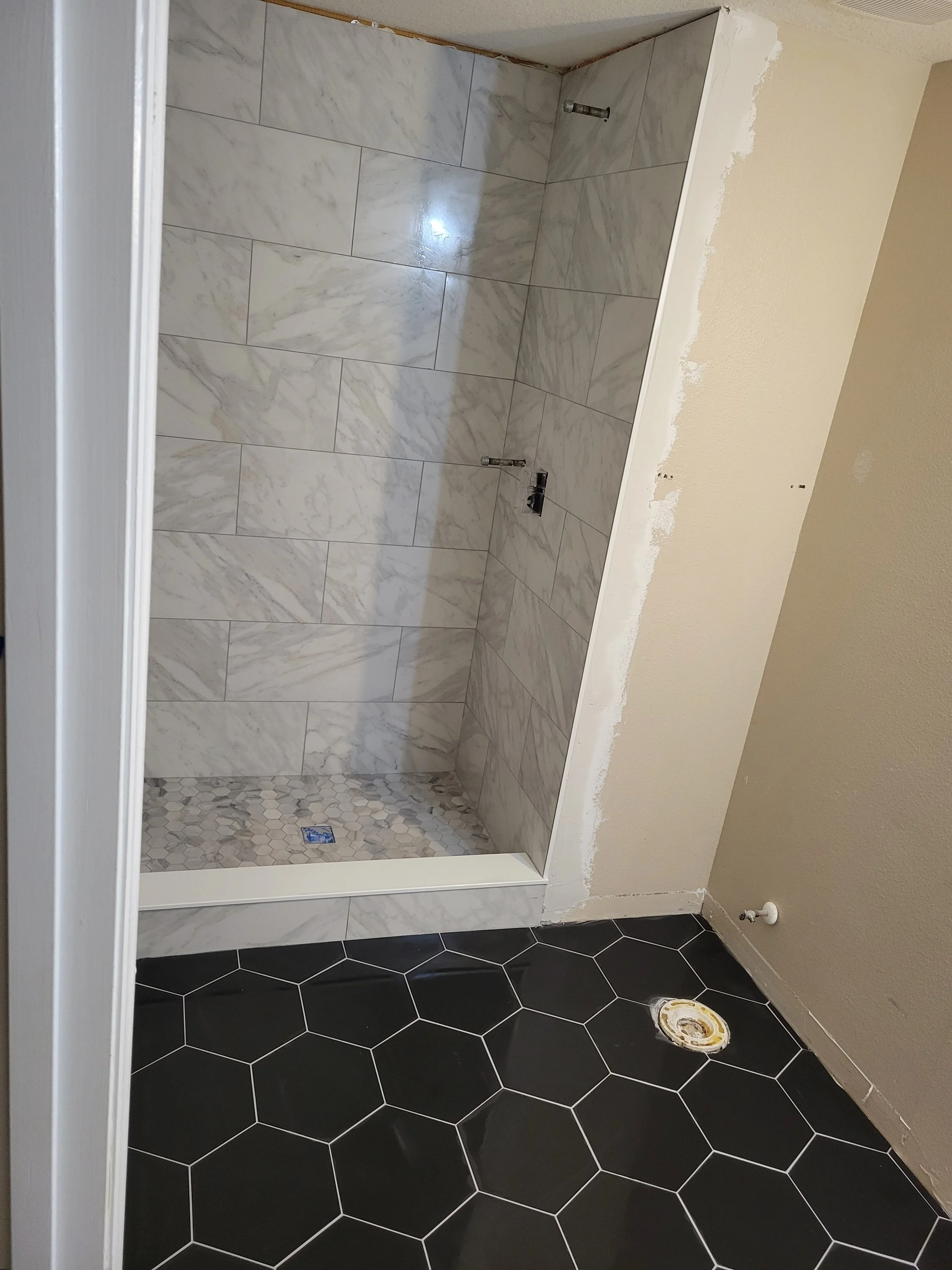 Bathroom shower area with white marble tiles on the walls and hexagonal black tiles on the floor, with plumbing fixtures and a drain in the corner.