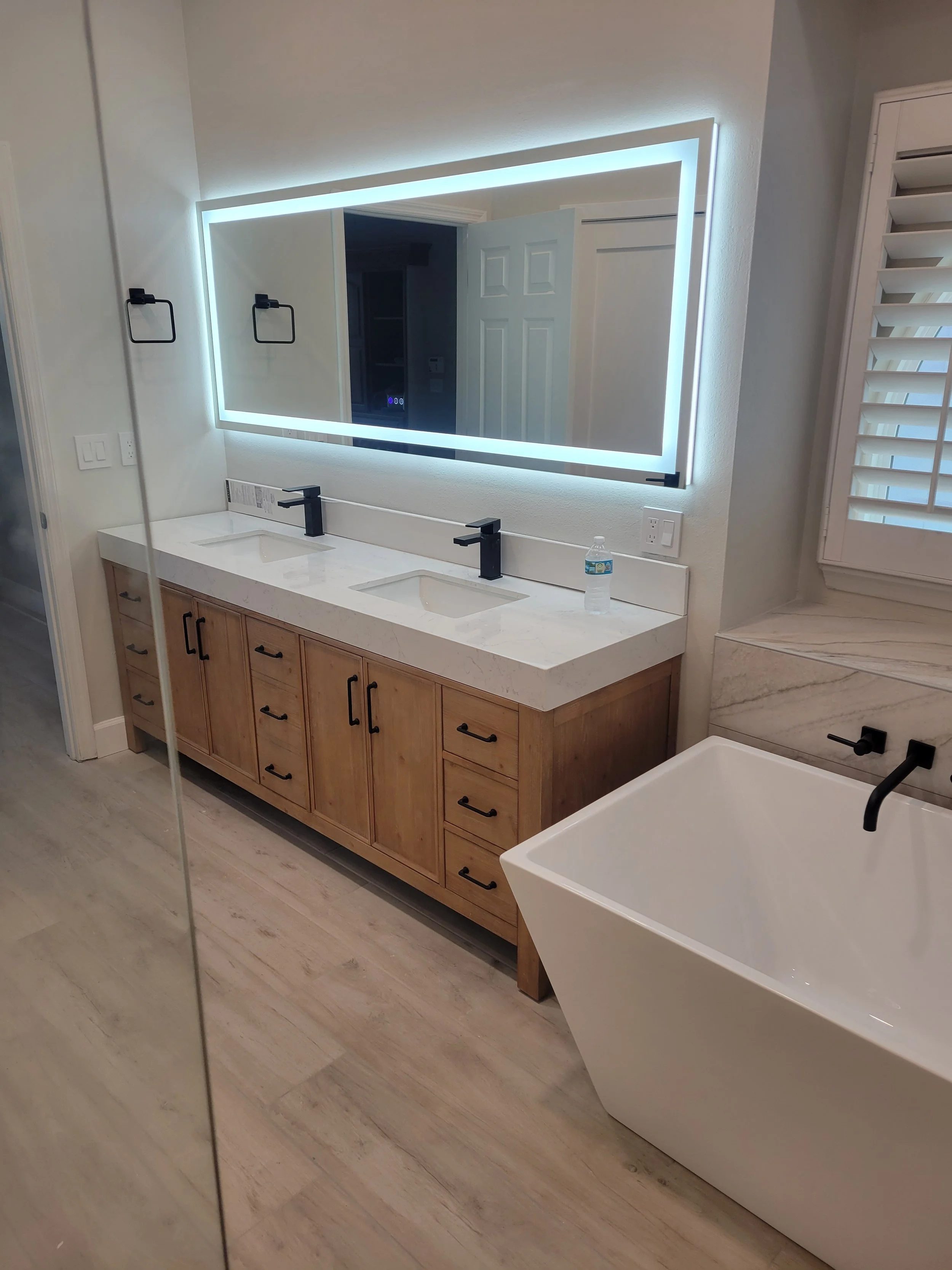 Modern bathroom with double vanity, large backlit mirror, freestanding bathtub, and light-colored wood flooring.