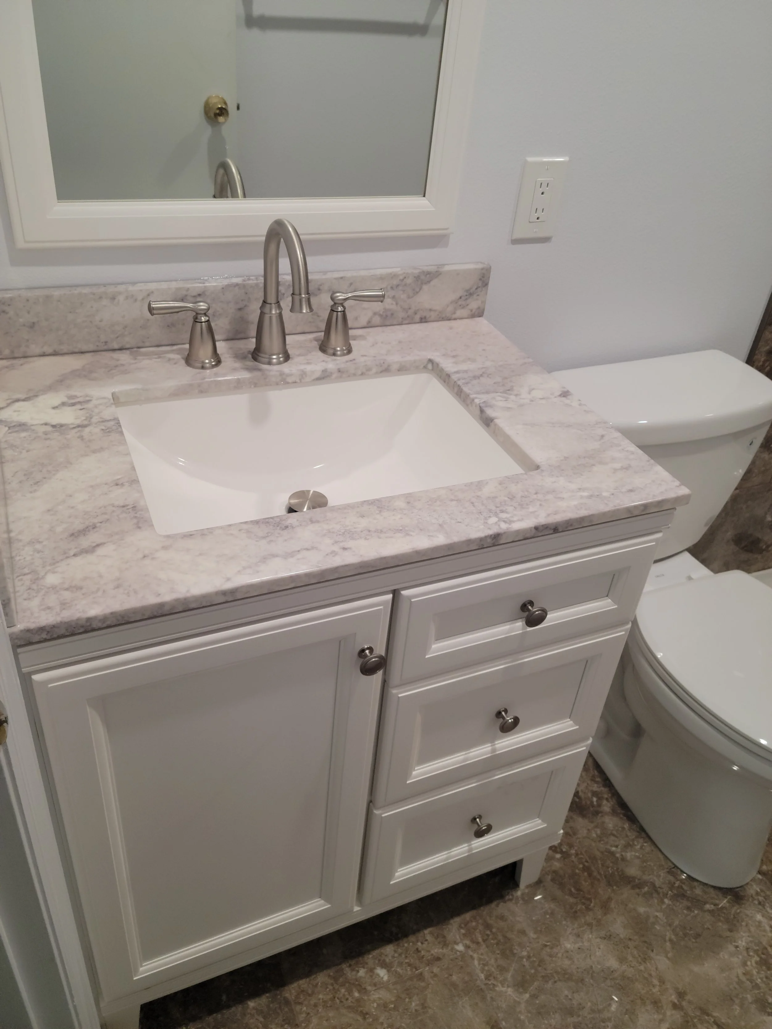 Bathroom vanity with a marble countertop, white undermount sink, silver faucet, and a mirror above. To the right, a white toilet.