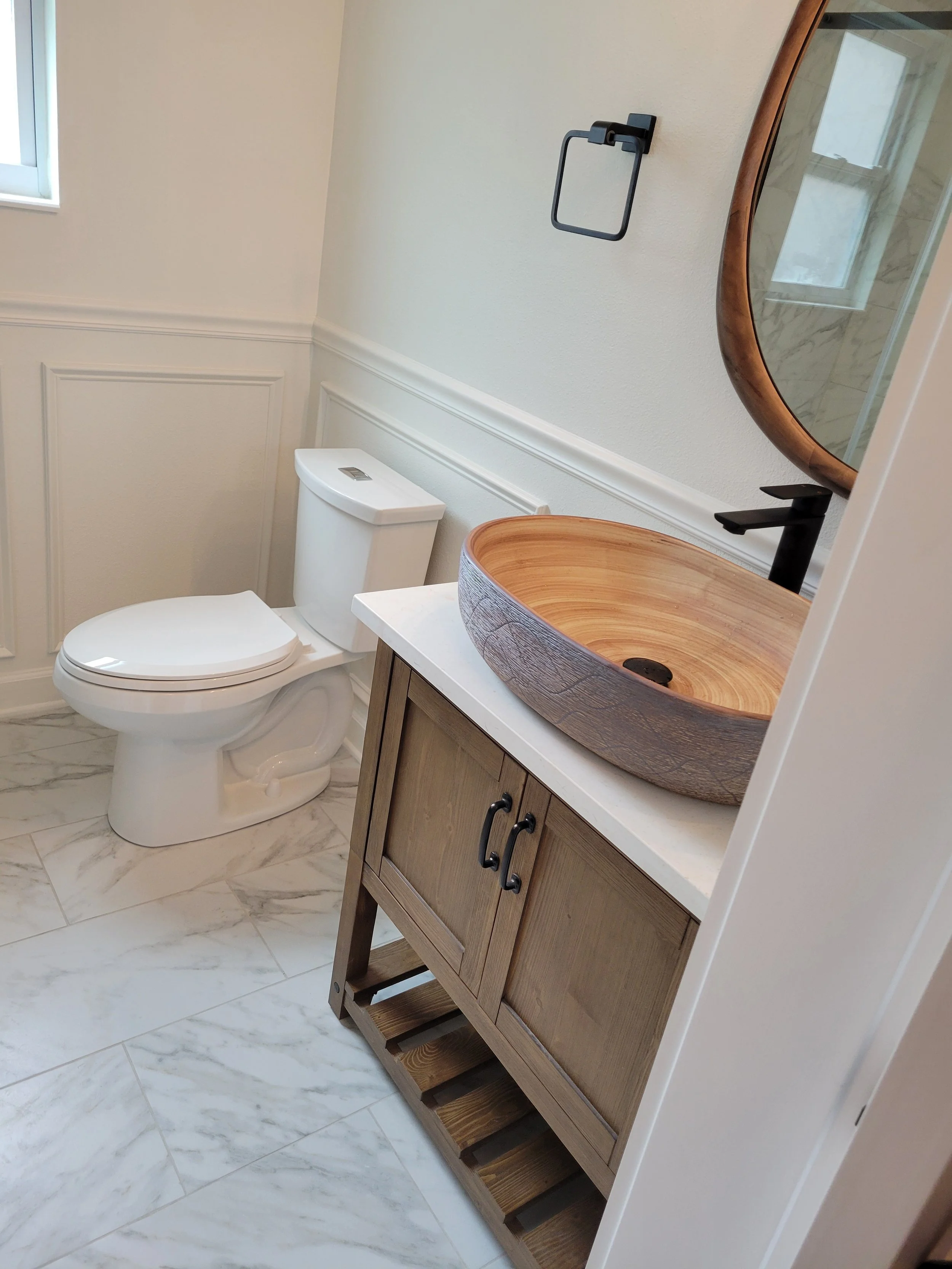 Bathroom with white toilet, wooden vanity with a vessel sink, oval mirror, black faucet, and marble floor tiles.