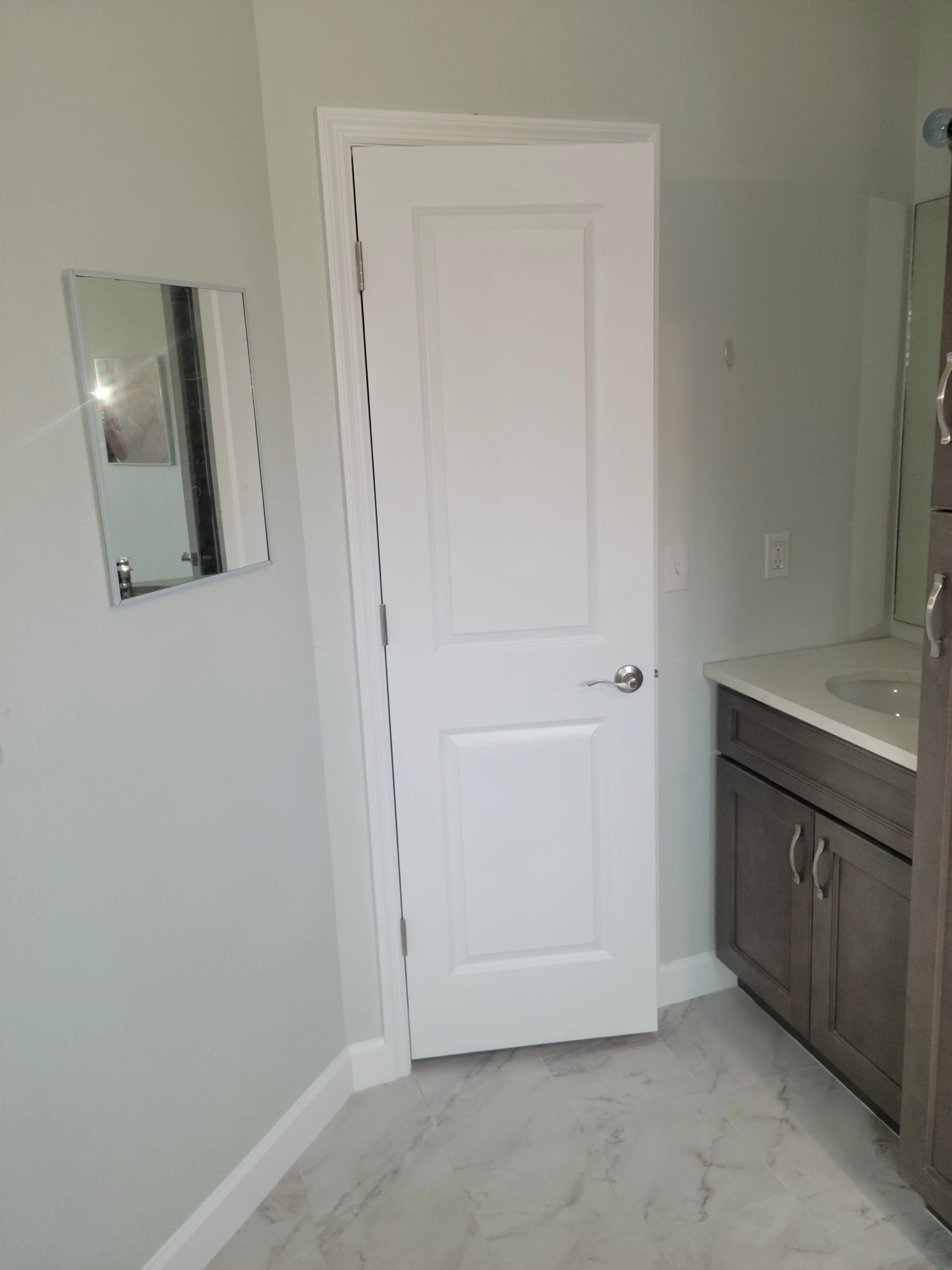 A white interior bathroom door beside a small mirror on a light-colored wall and a bathroom vanity with gray cabinet doors and a white countertop.
