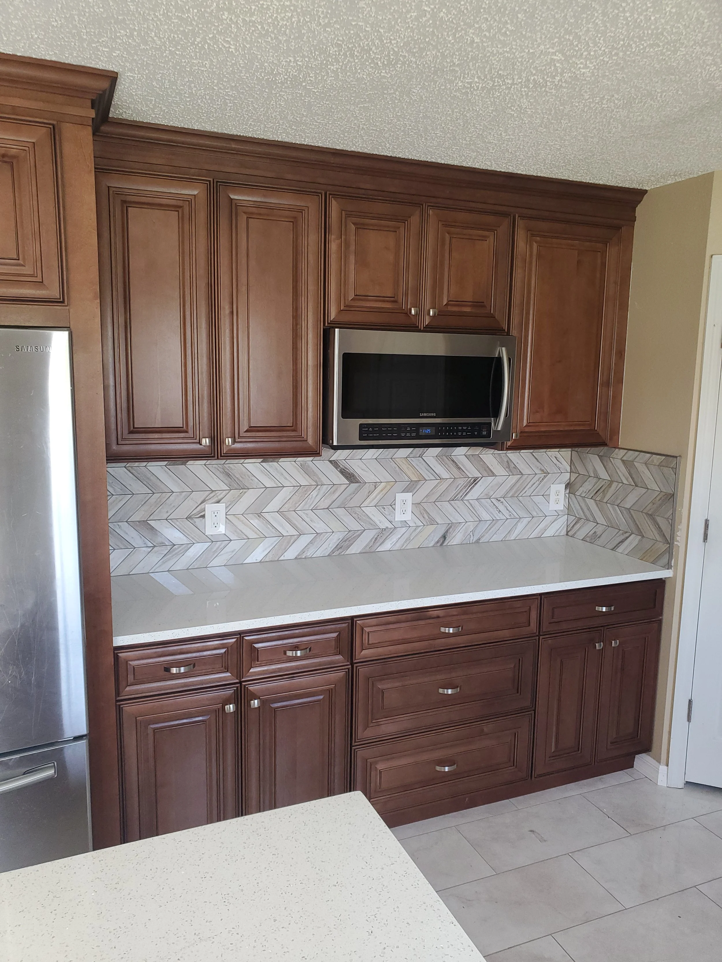 Kitchen with wooden cabinets, a microwave, a white countertop, a stainless steel refrigerator, and a textured tile backsplash.