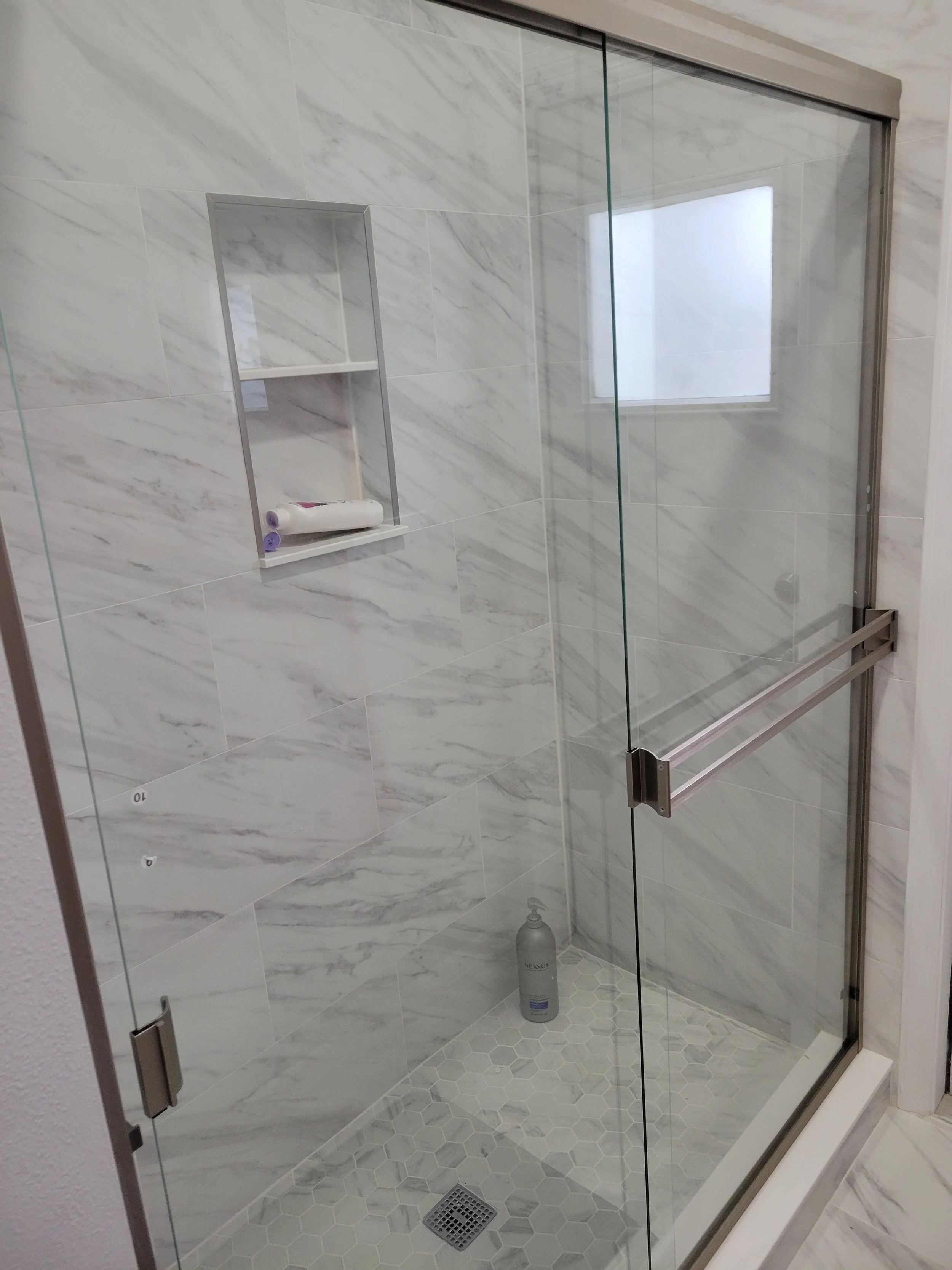 A modern glass shower enclosure with marble wall tiles, a built-in shelf with toiletries, and a small window.