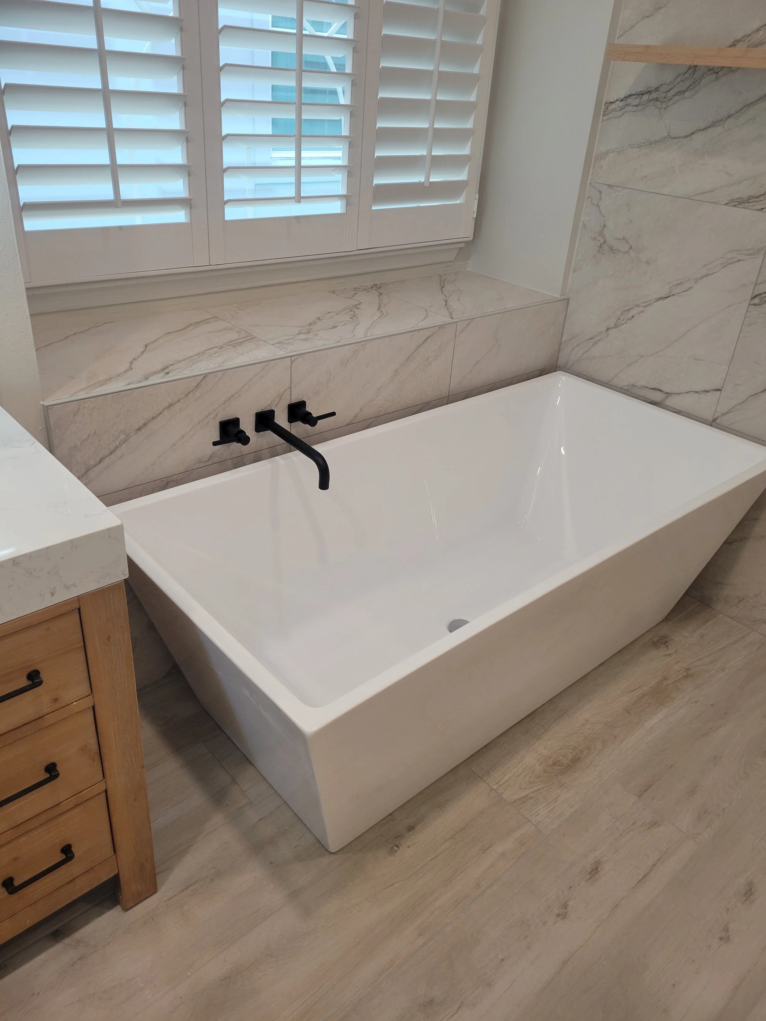 White modern bathtub with black faucet and handles in a bathroom with marble tiles and a window with white shutters.
