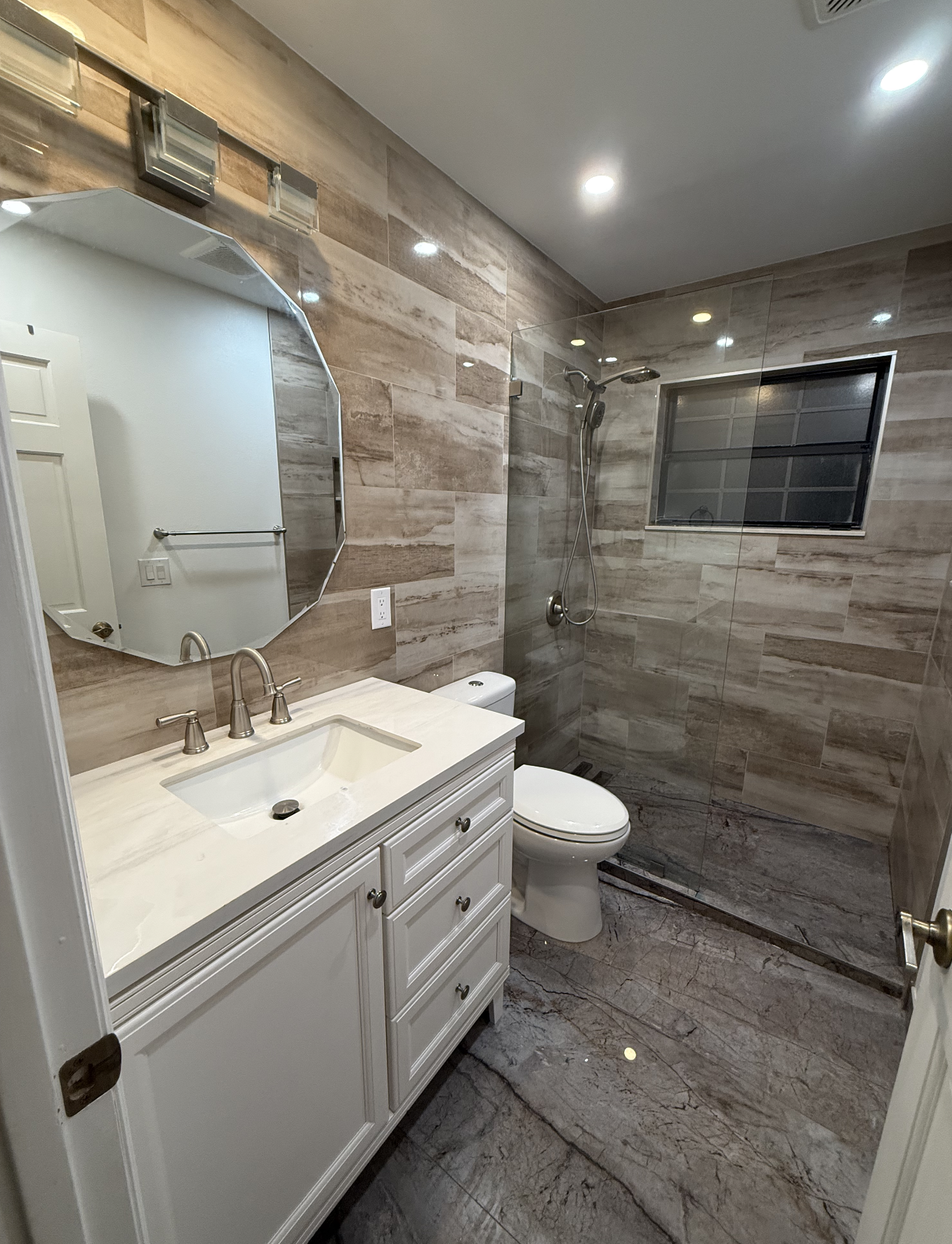 A modern bathroom with a white vanity and marble countertop, a large mirror, a toilet, and a walk-in shower with beige marble tiles, a small window, and a glass partition.