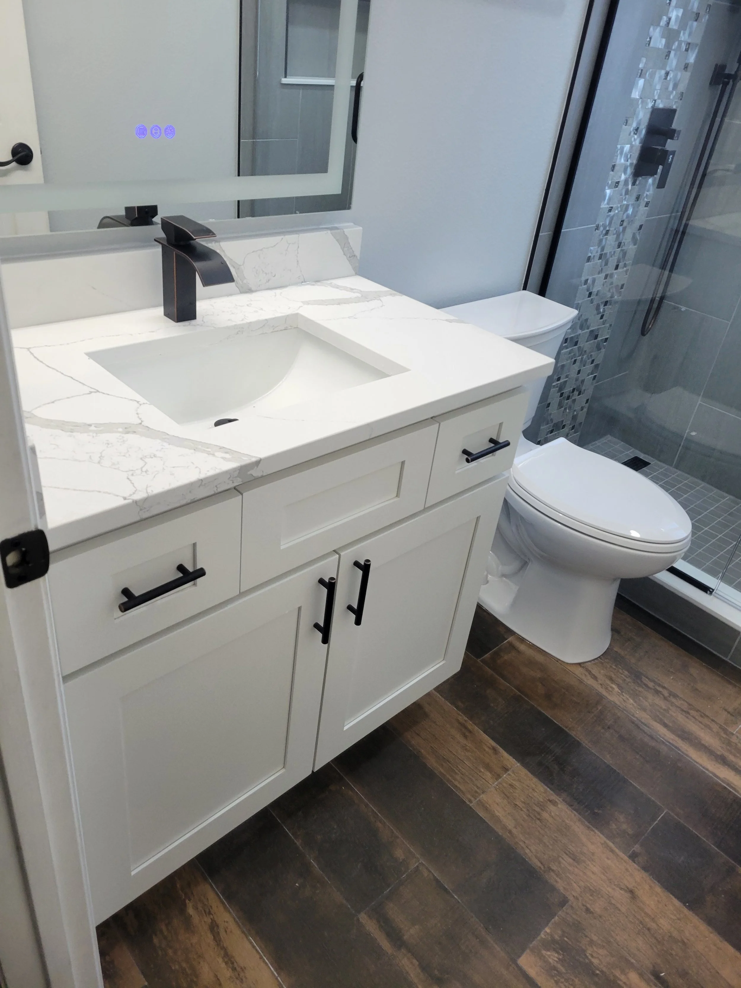 Modern bathroom with white vanity featuring a marble countertop, black faucet, and a large mirror. To the right, a white toilet is next to a glass-enclosed shower with gray tiles and a mosaic accent wall.