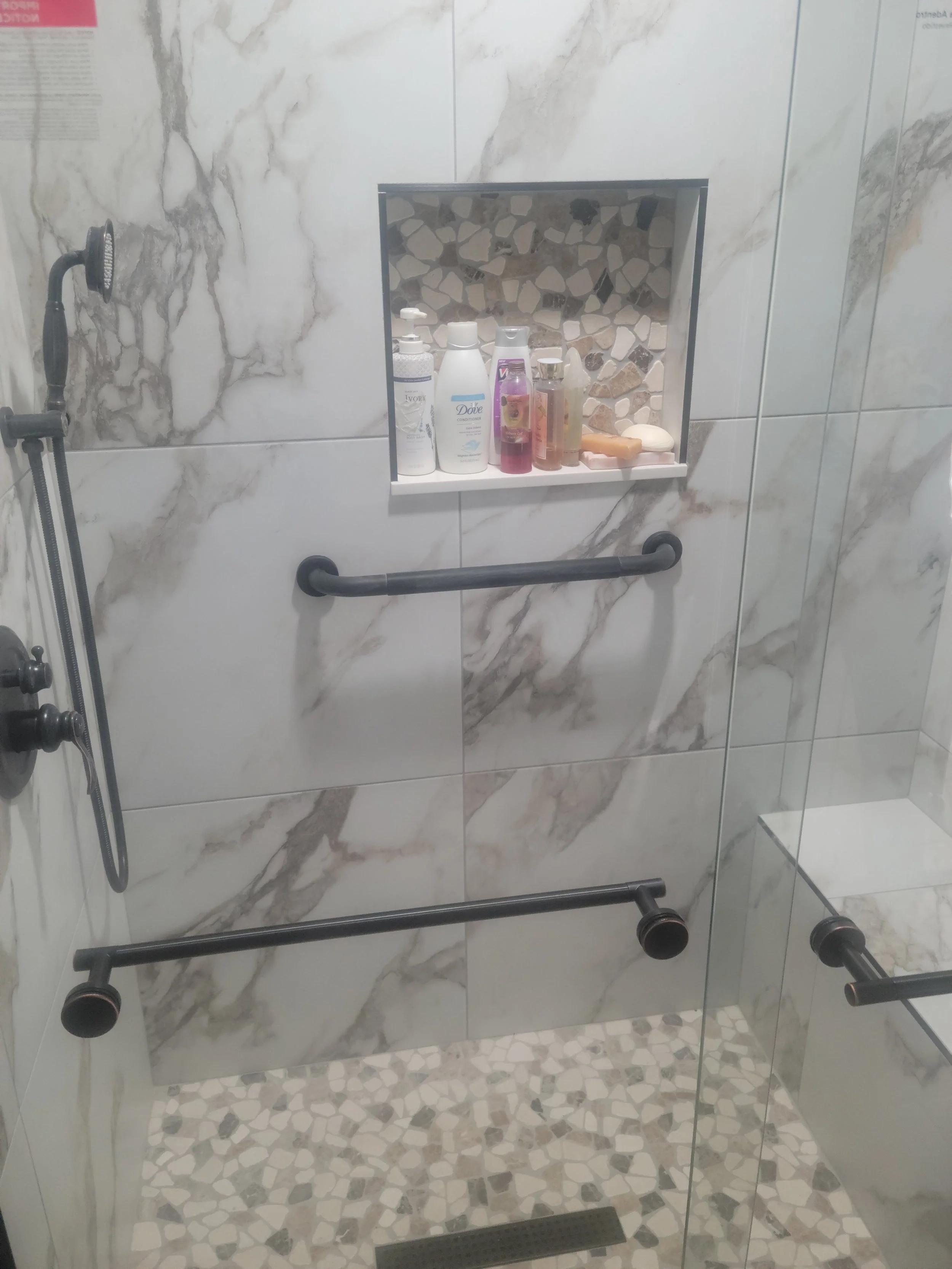 Shower with marble tiles, a built-in shelf containing bottles of shampoo, soap, and lotion, black grab bars, a hand-held shower head, and a glass door.