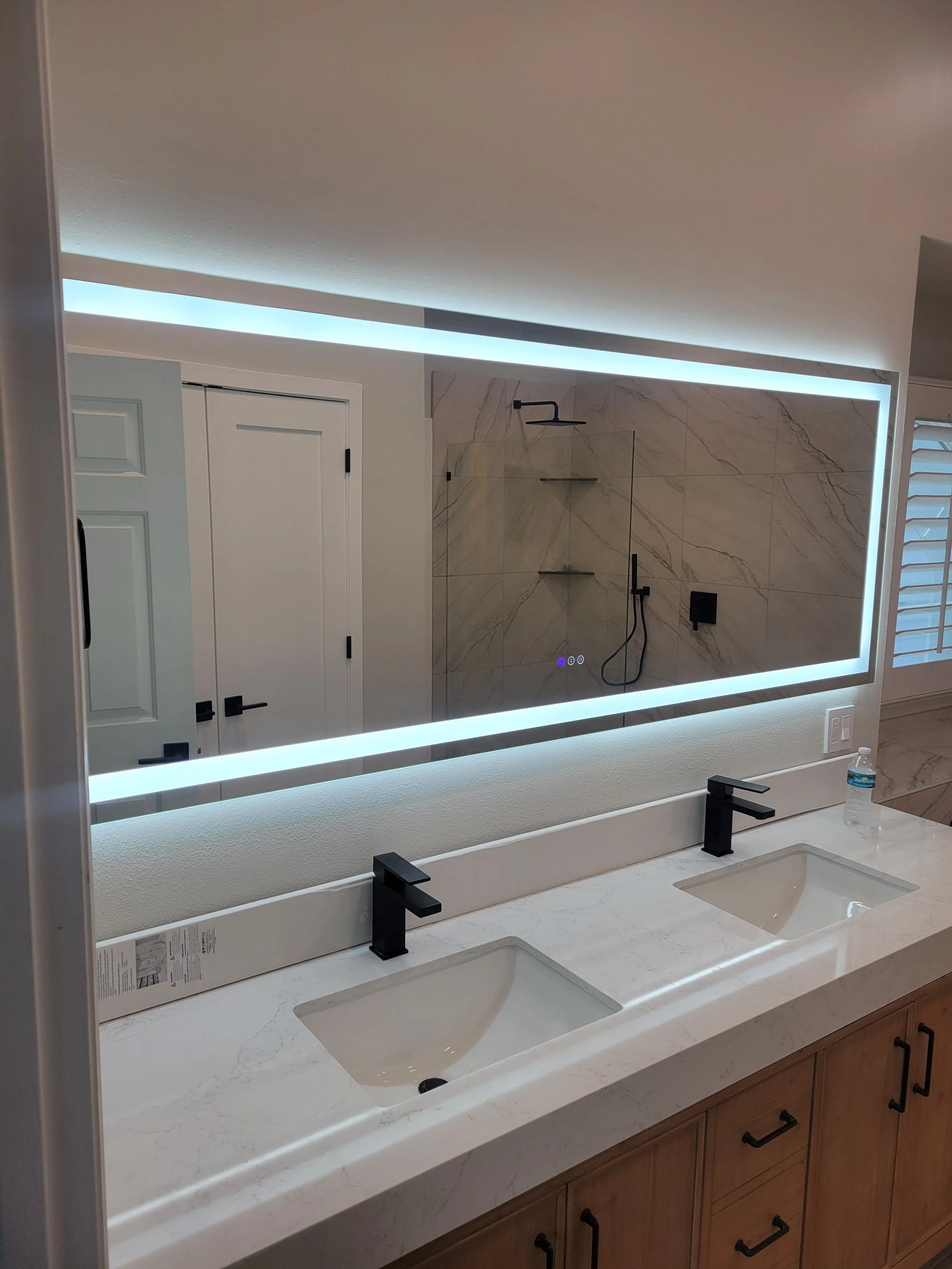 Modern bathroom with dual sinks, black fixtures, a large illuminated mirror, and a walk-in shower with marble tiles.
