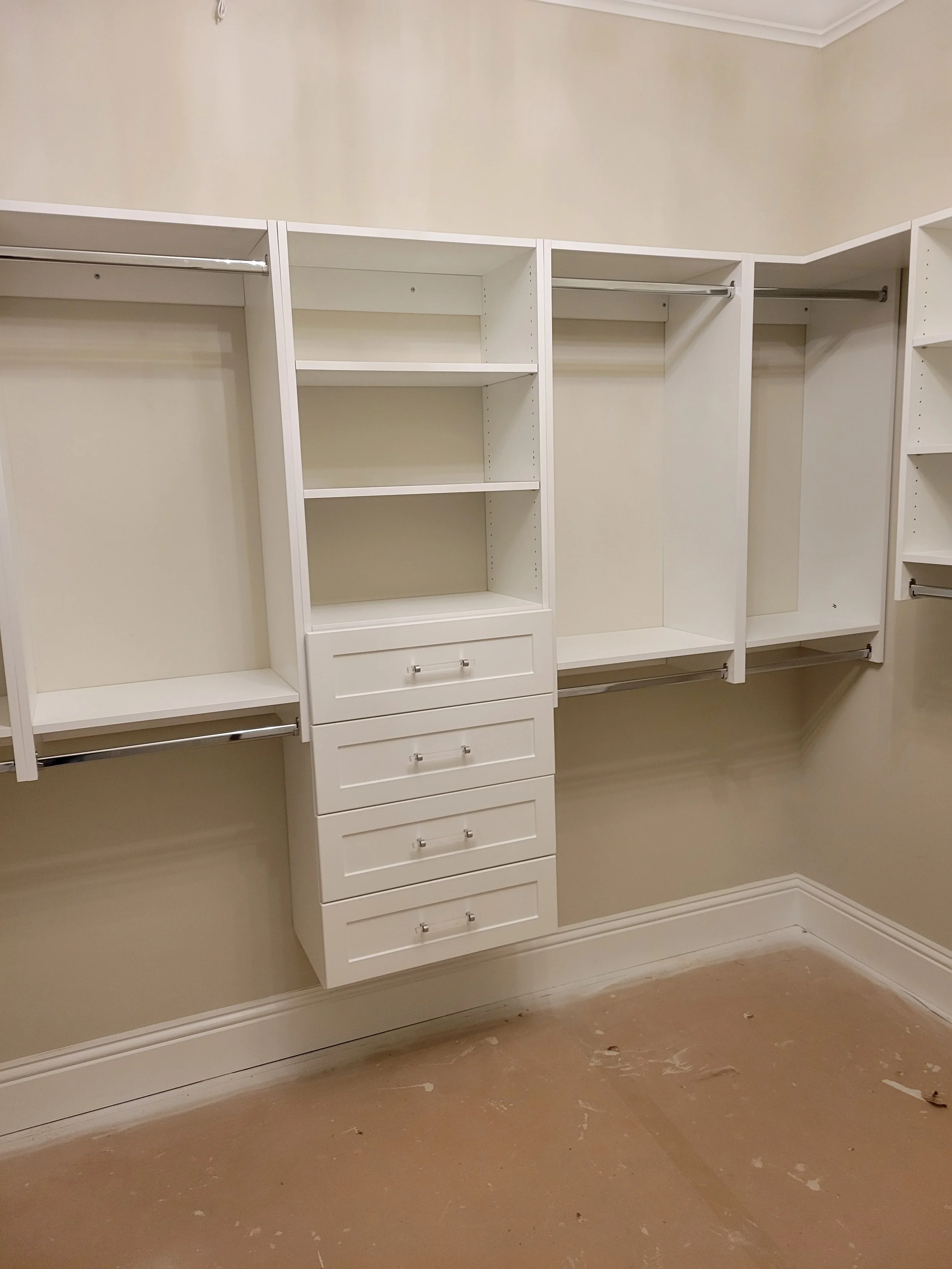 Empty walk-in closet with built-in white shelving and hanging rods, beige walls, and a unfinished floor.