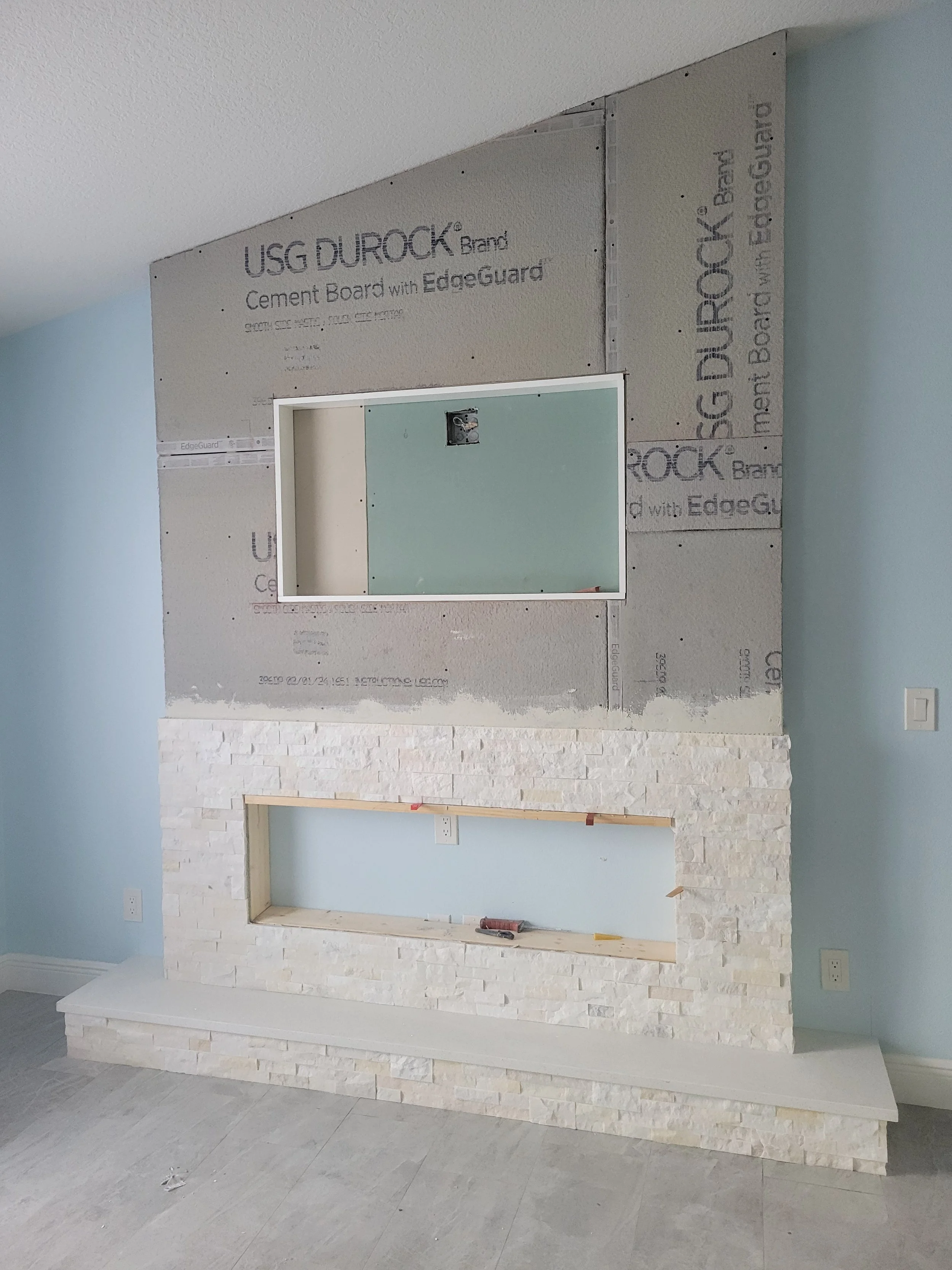 Partially built fireplace with stonework and an opening framed for a mantel, in a room under construction, with blue walls and a wall with installed cement board.