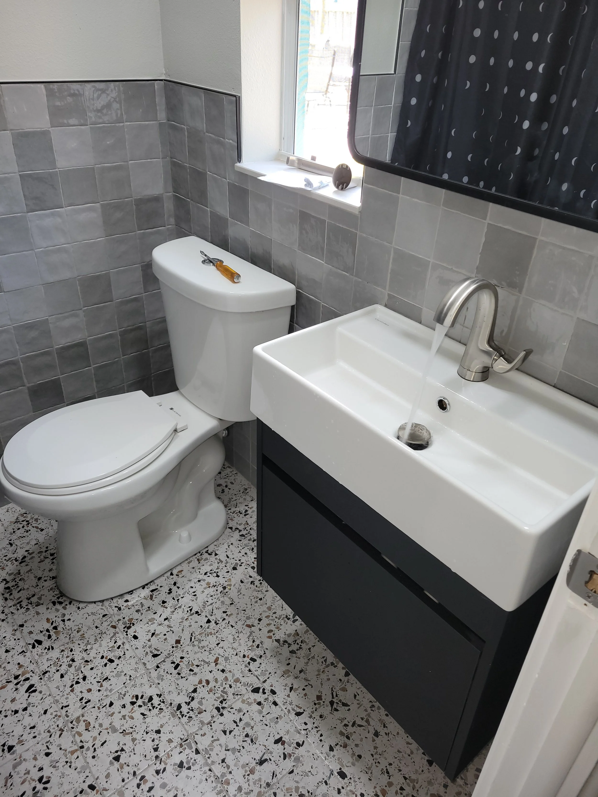 Bathroom with a toilet, small sink with a running faucet, and a window with a polka dot shower curtain in the background.