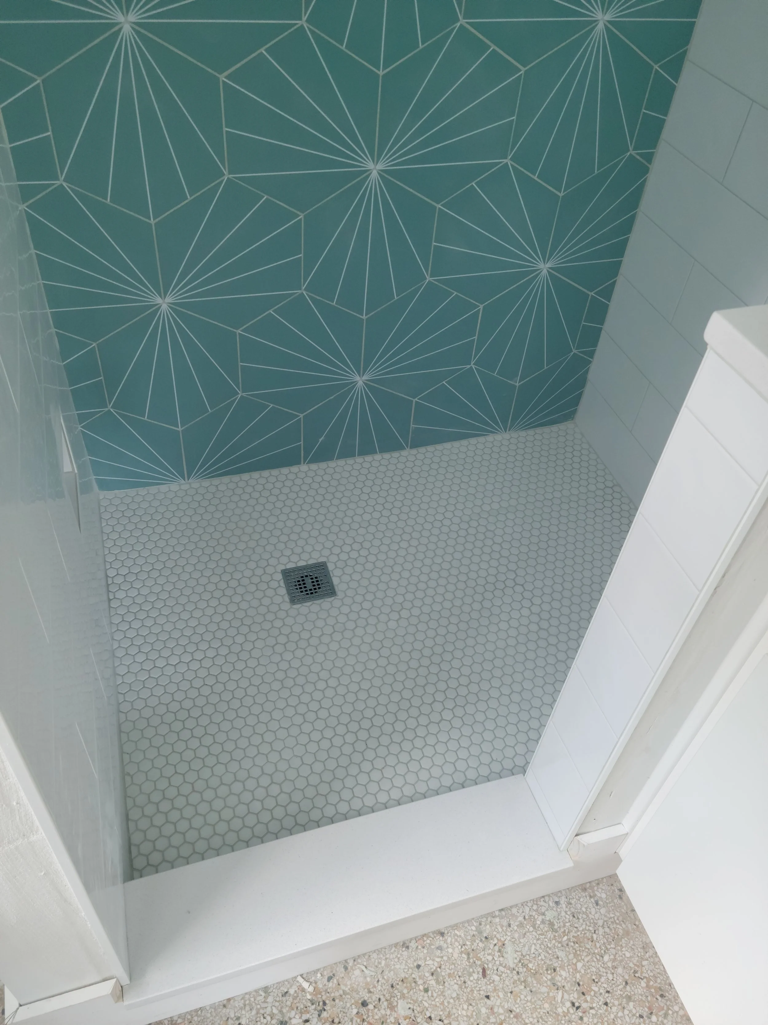 Shower area with white hexagonal tile floor, teal wall with geometric line art pattern, and a metal drain cover.