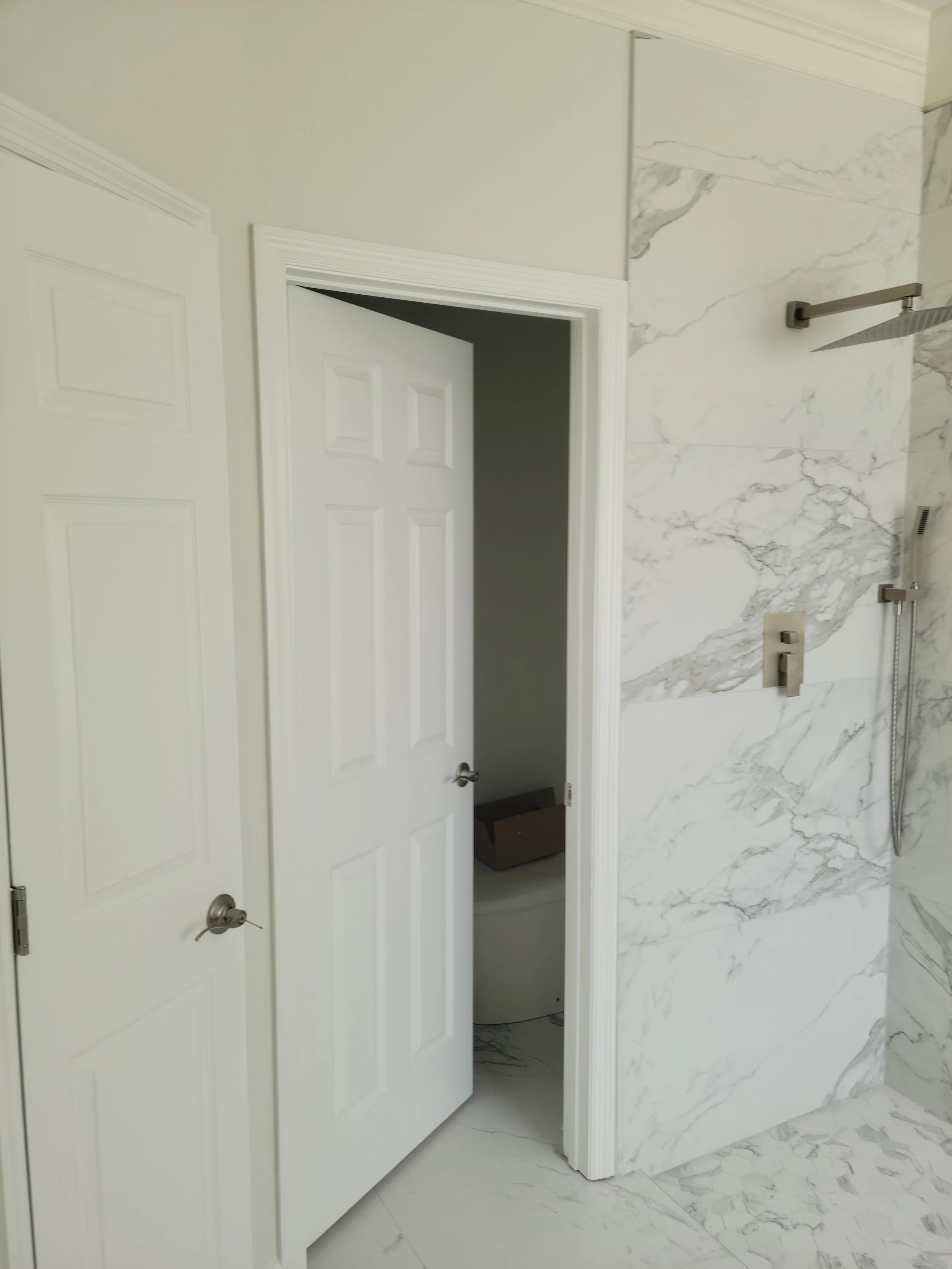 A bathroom with a marble shower wall, a partially open white door revealing a toilet inside, and another closed door to the left.