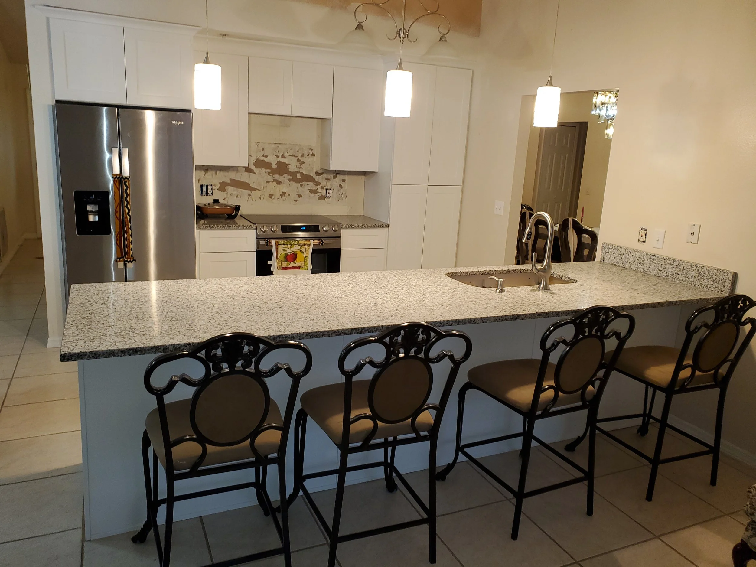 Kitchen with a granite island counter, four bar stools, white cabinets, stainless steel refrigerator, stove, and pendant lights.