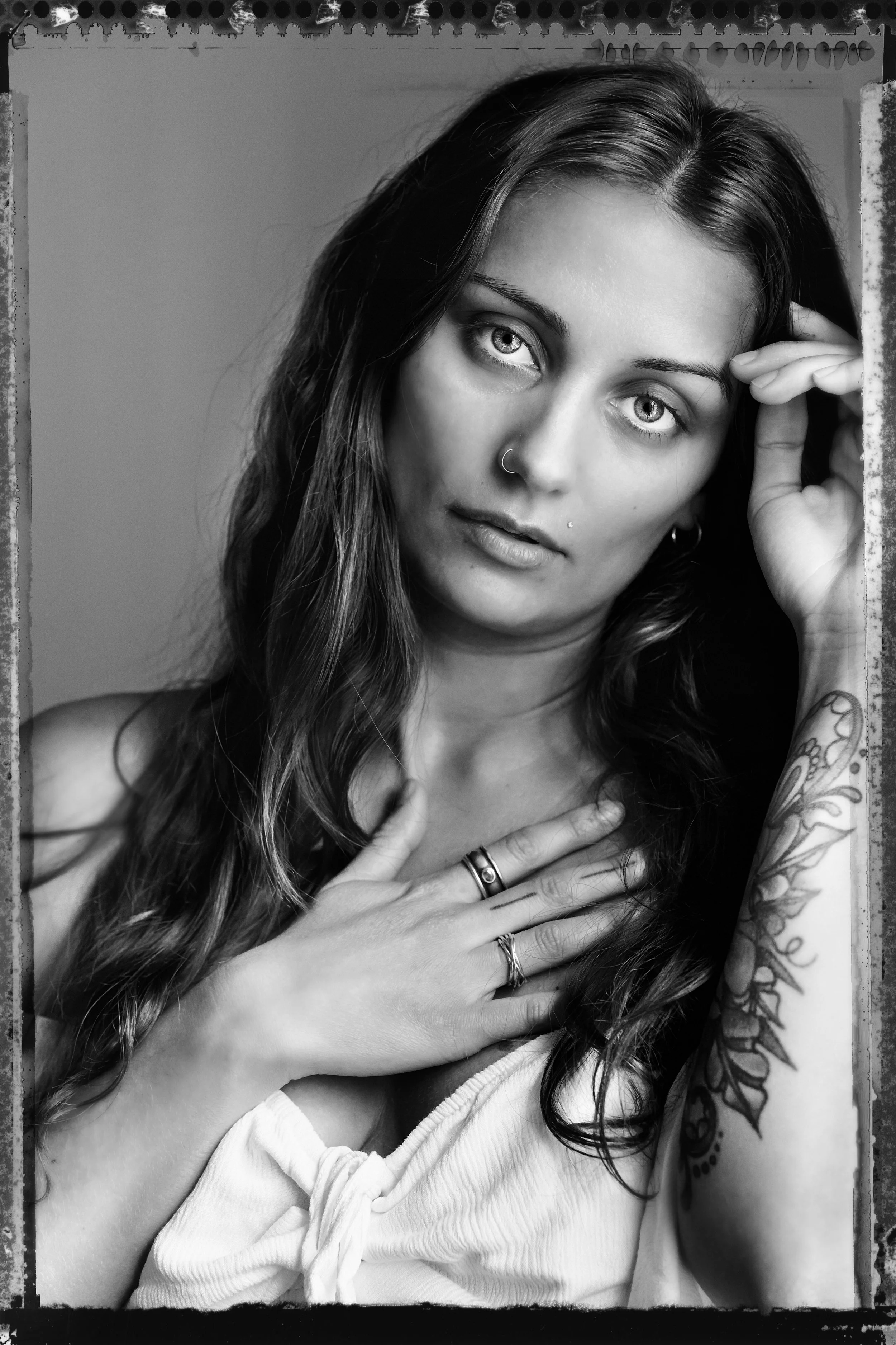 Black and white portrait of a woman with long hair, a nose ring, tattoo on her arm, hand on her chest, and wearing a sleeveless top.