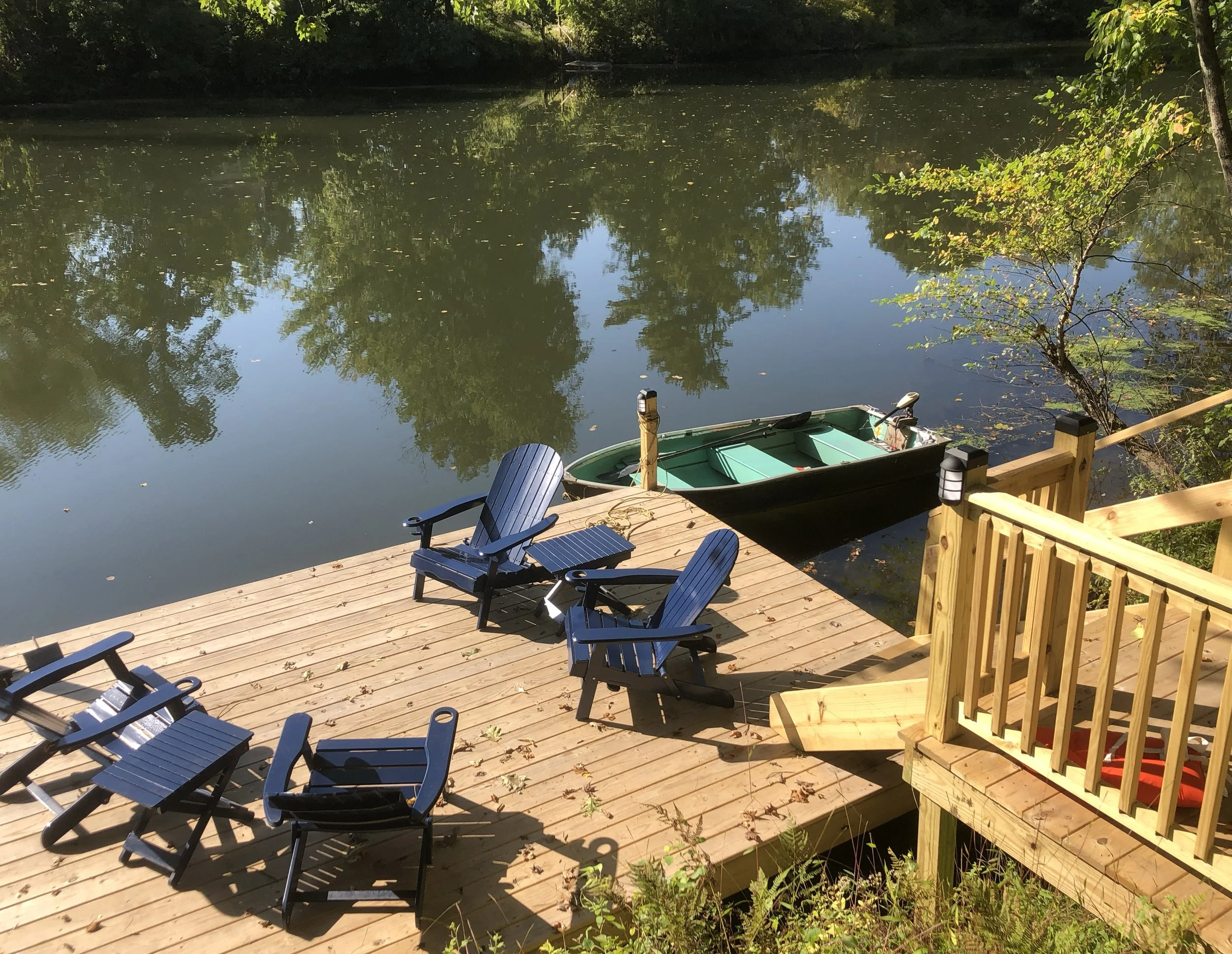 Wooden dock with Adirondack chairs next to a calm river; a small boat is moored at the dock.