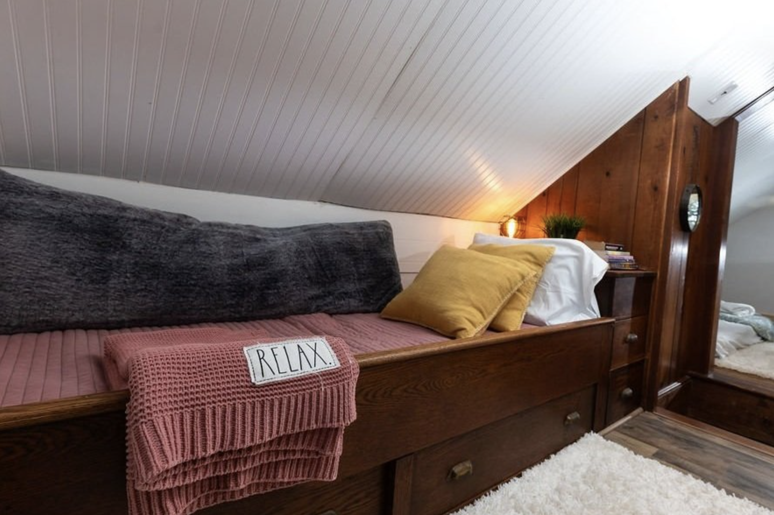 Cozy attic bedroom with wooden bed, yellow and grey pillows, pink blanket labeled 'RELAX', and a lamp on a bedside table with books and a plant.