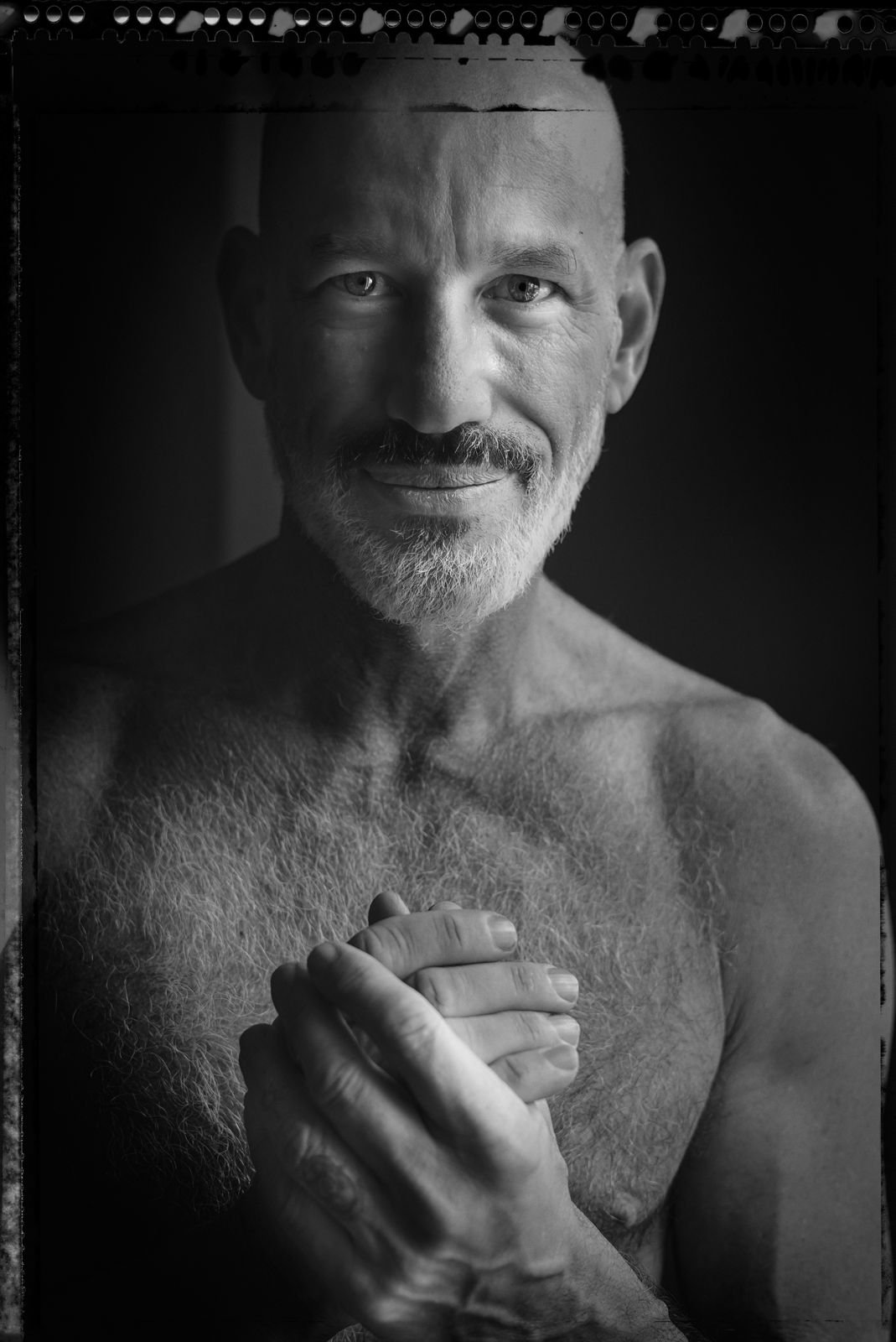 Black and white portrait of a shirtless man with a beard, smiling and holding his hands together.