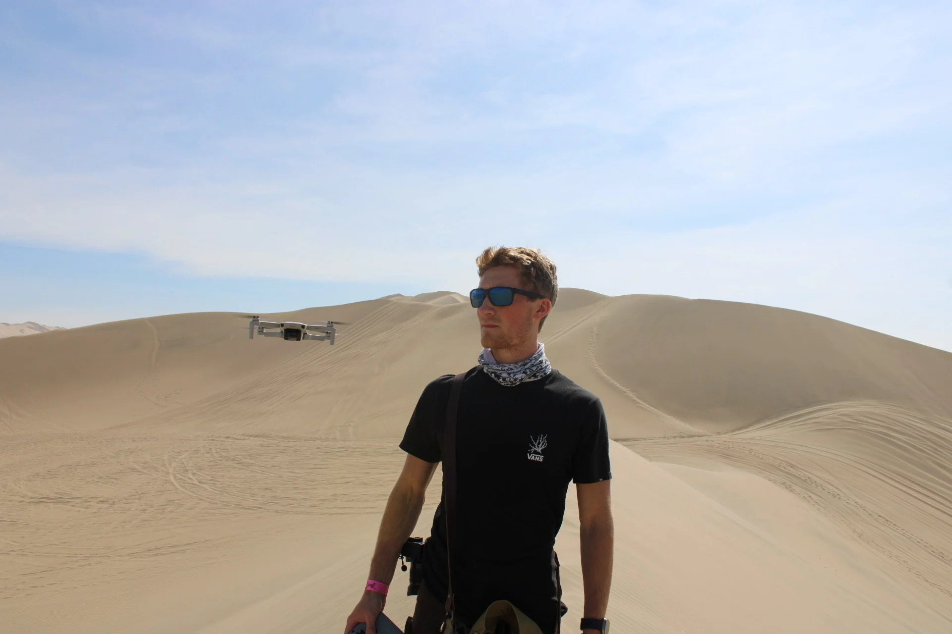 A man wearing sunglasses and a black t-shirt with a logo stands in a desert with sand dunes and a drone flying nearby.