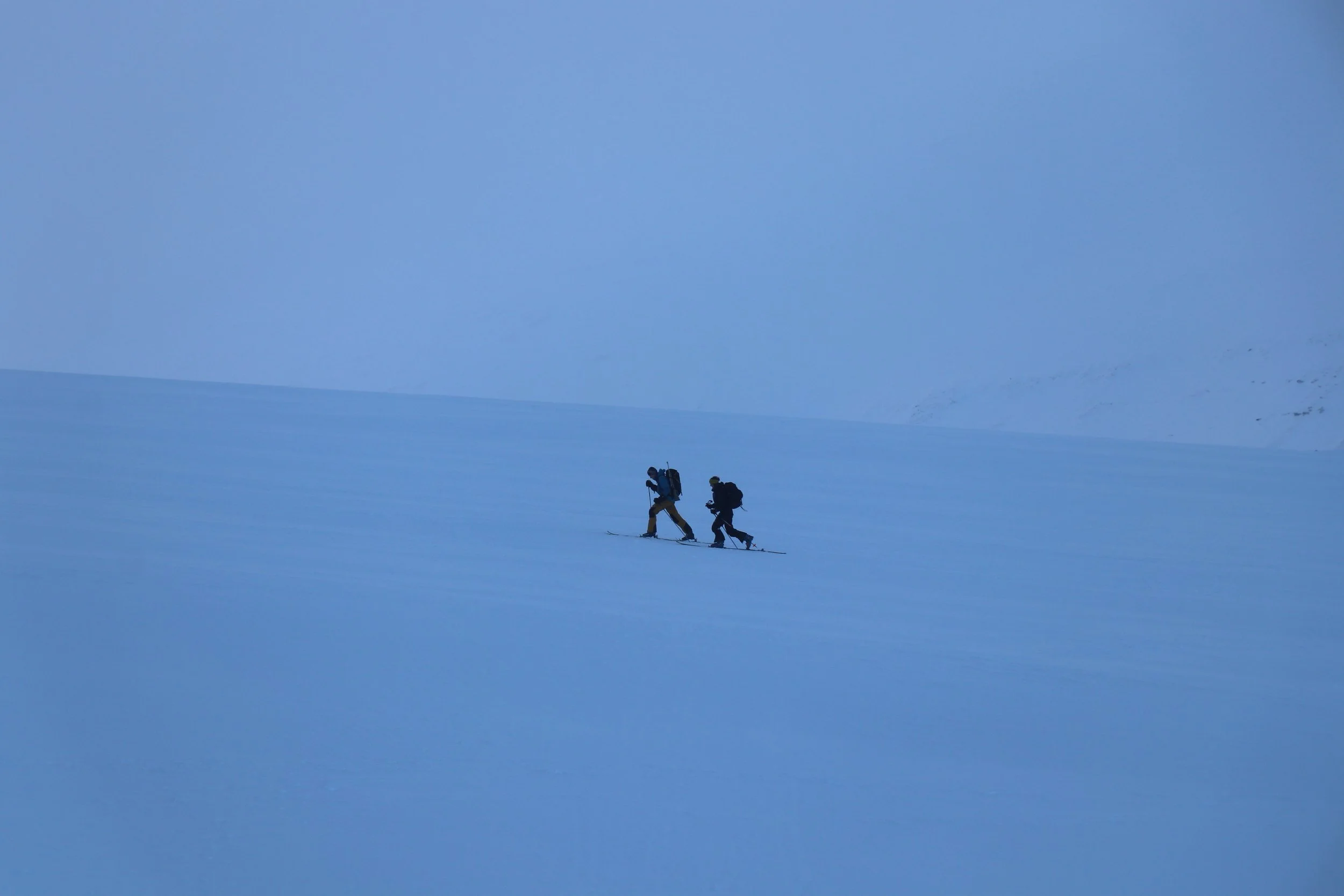 Two people cross-country skiing on a snow-covered landscape with a mountain in the background.