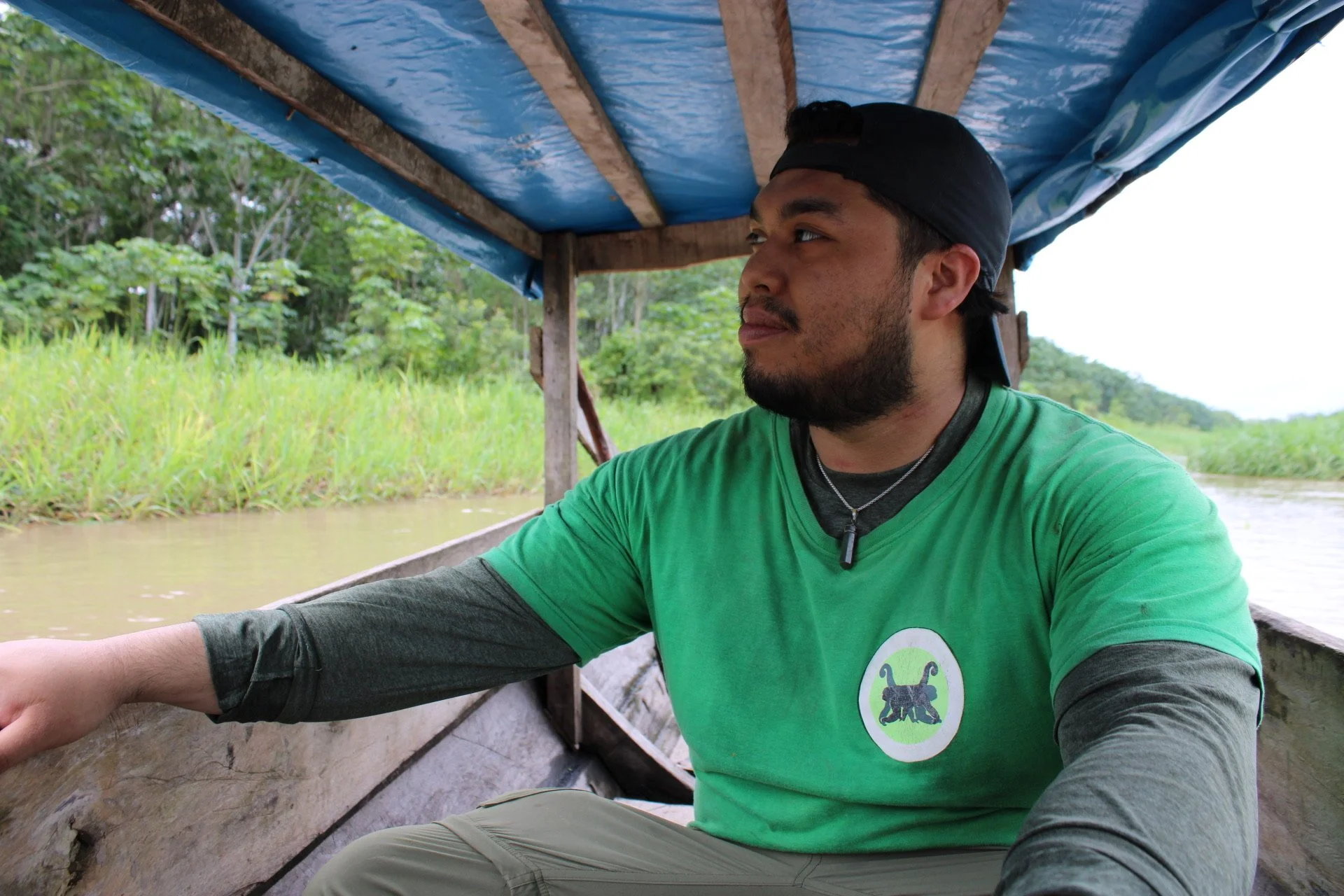 Man sitting in a wooden boat on a river, wearing a green shirt and a black cap, with lush green trees and grass in the background.