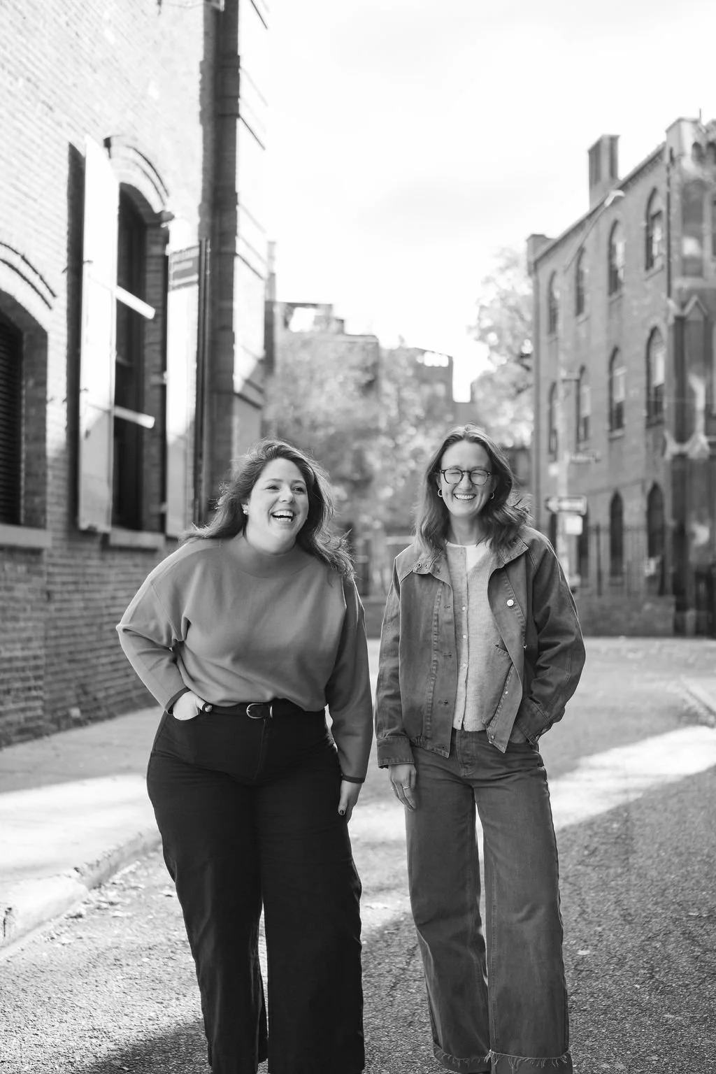 Two women laughing and walking on a street with brick buildings, in black and white.