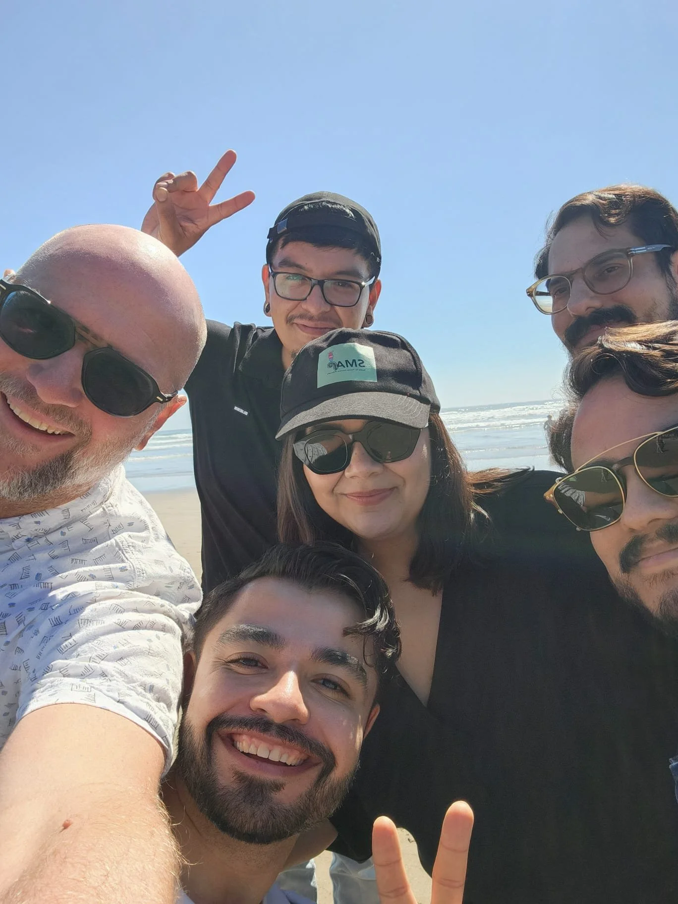 Group of friends taking a selfie on the beach with ocean waves in the background, smiling and making peace signs.