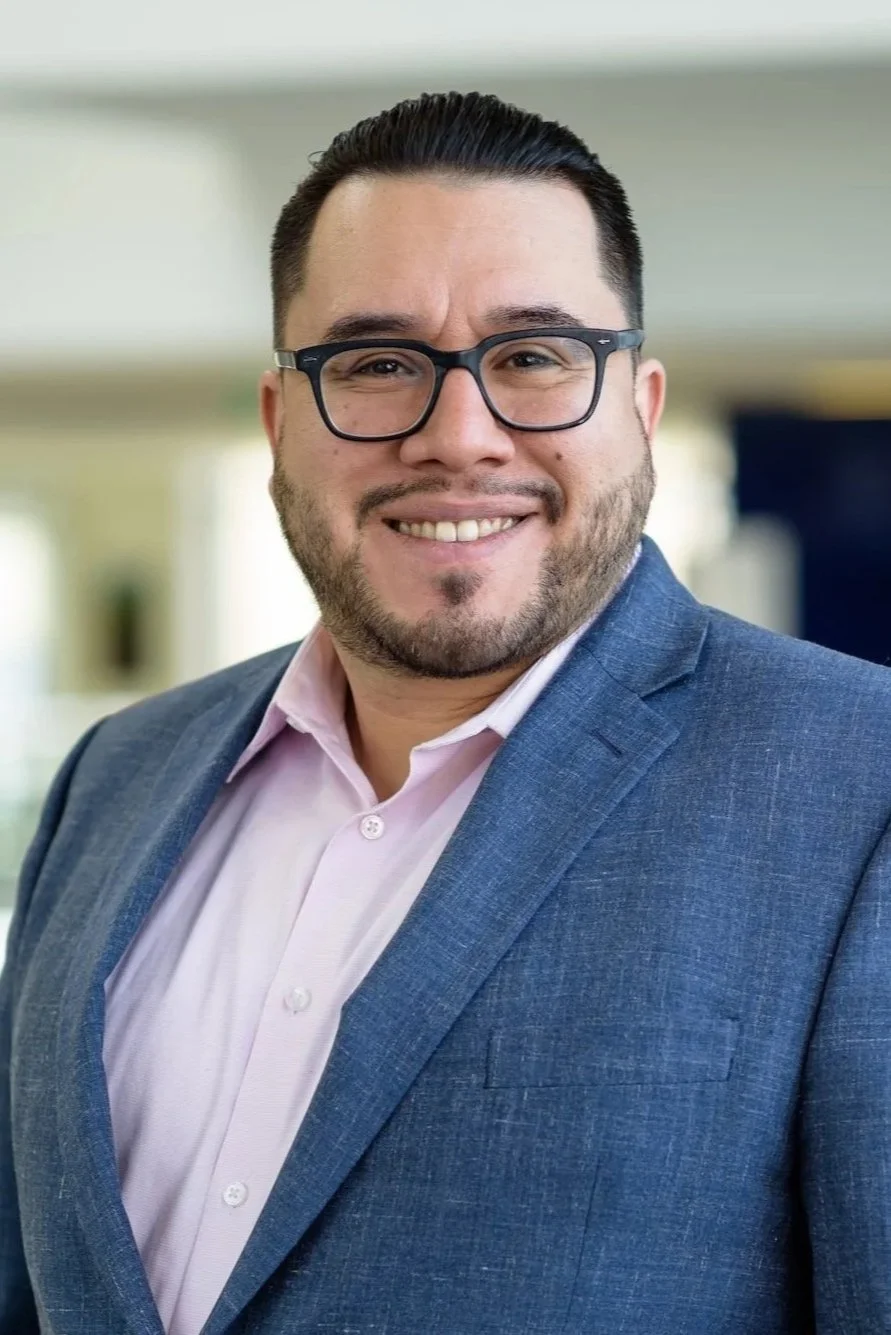 A smiling man with black hair, glasses, and a beard, wearing a blue suit and pink shirt in an indoor professional setting.