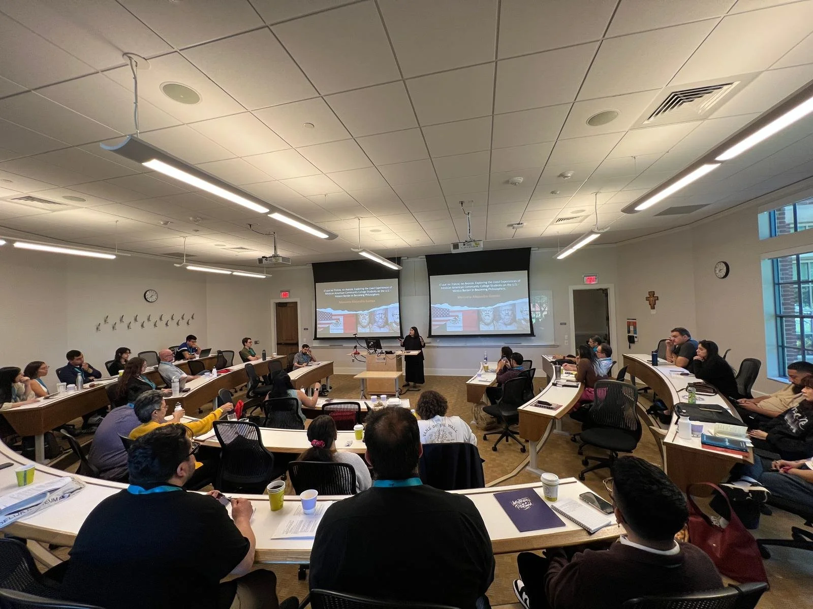 A classroom or conference room filled with people seated at desks, attending a presentation with two large screens displaying a PowerPoint slide. The room has white walls, two clocks, a crucifix, large windows on the right, and a woman standing at a 