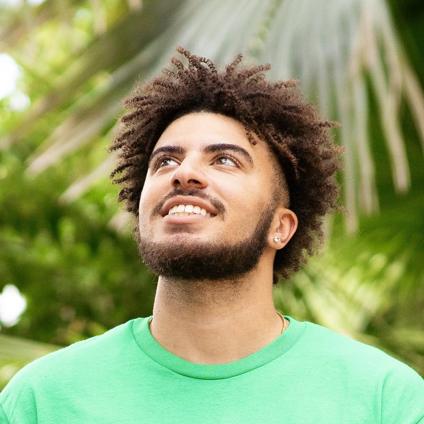 Man in green shirt smiling and looking up, standing in front of green foliage.