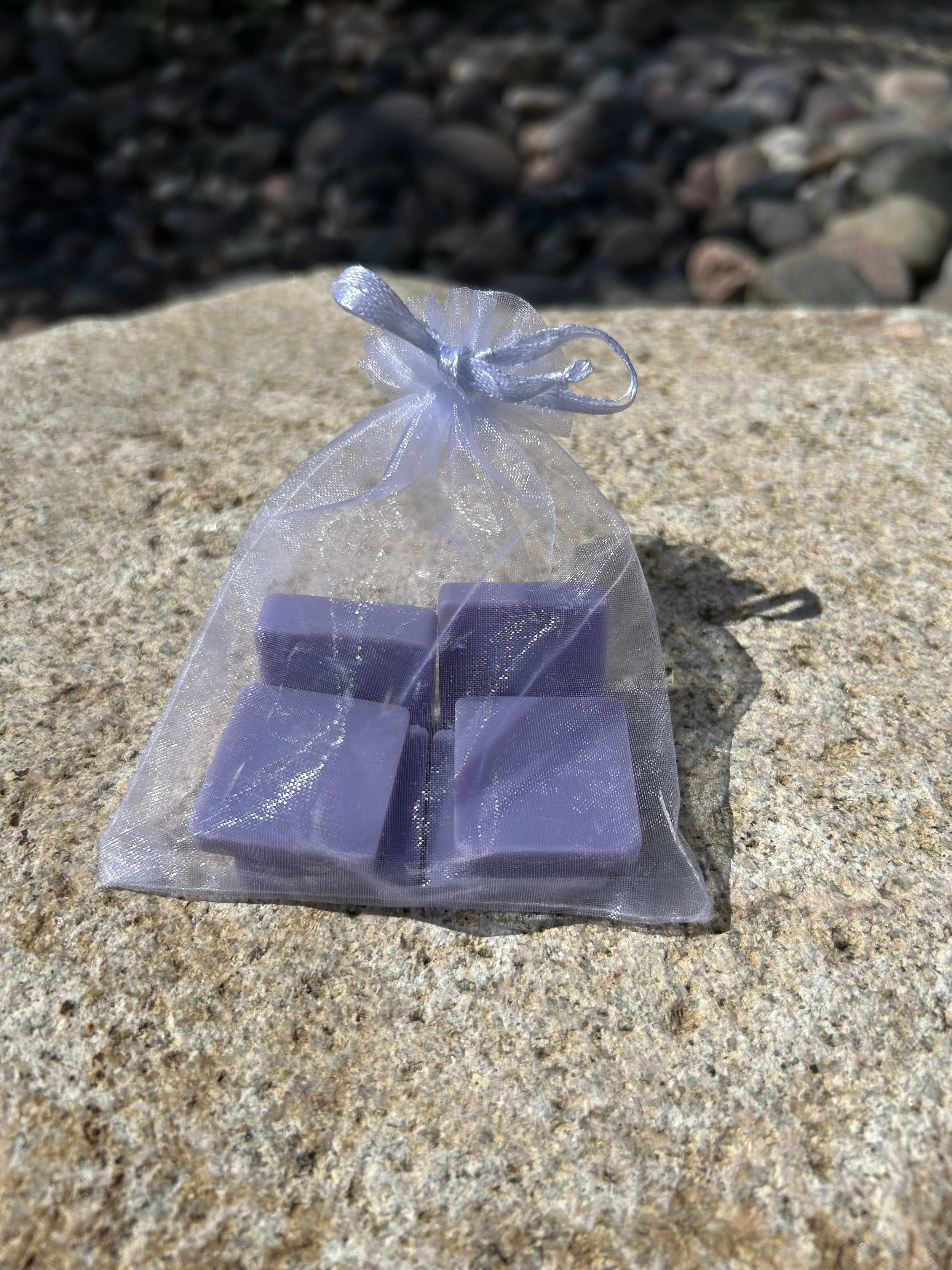 A small organza gift bag with a ribbon bow on top, with purple lavender scented wax melts inside. It's placed on a large stone surface with rocks in the background.