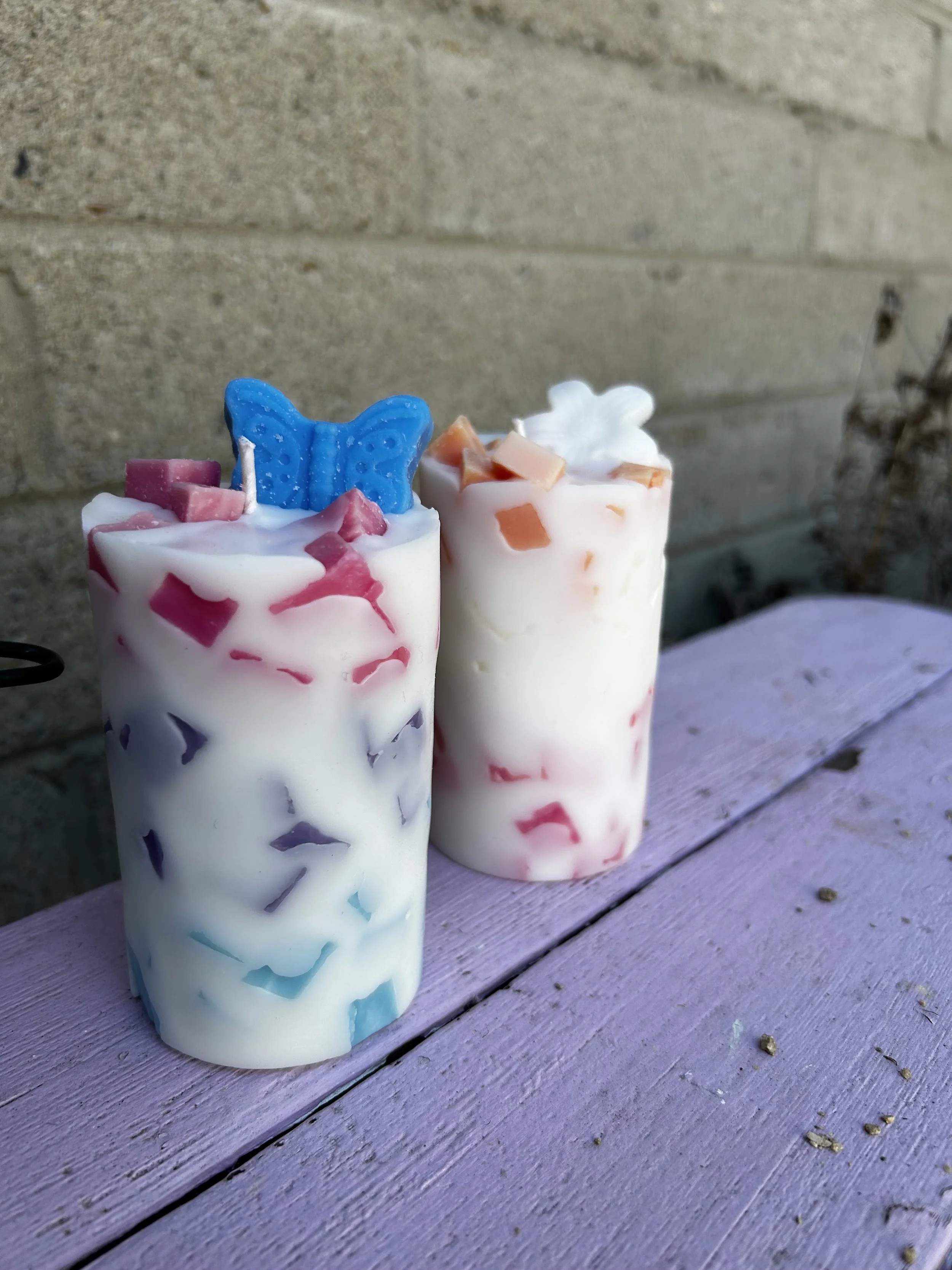 Two candles with spring and pastel colors, topped with a butterfly and flower topper decoration. They are displayed outdoors on a light purple bench.